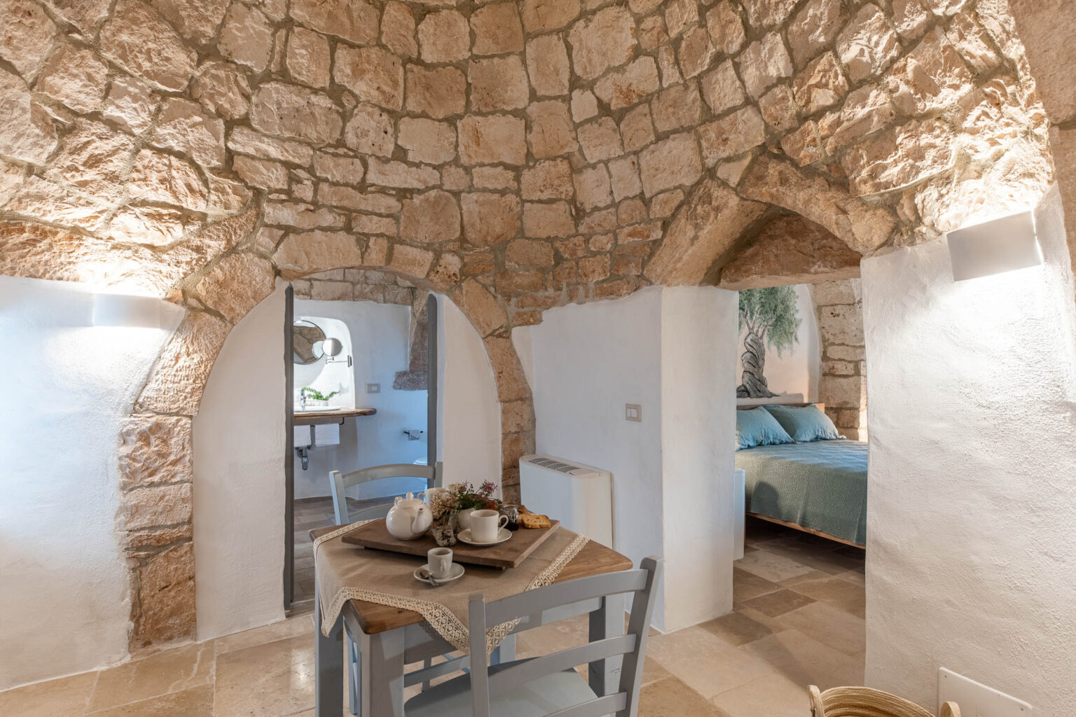 Charming interior of a rustic stone cottage featuring arched doorways, a cozy dining area with a wooden table and tea set, and a softly lit bedroom with a textured wall. The warm stone walls and modern decor create an inviting atmosphere. | Only on SopranoVillas
