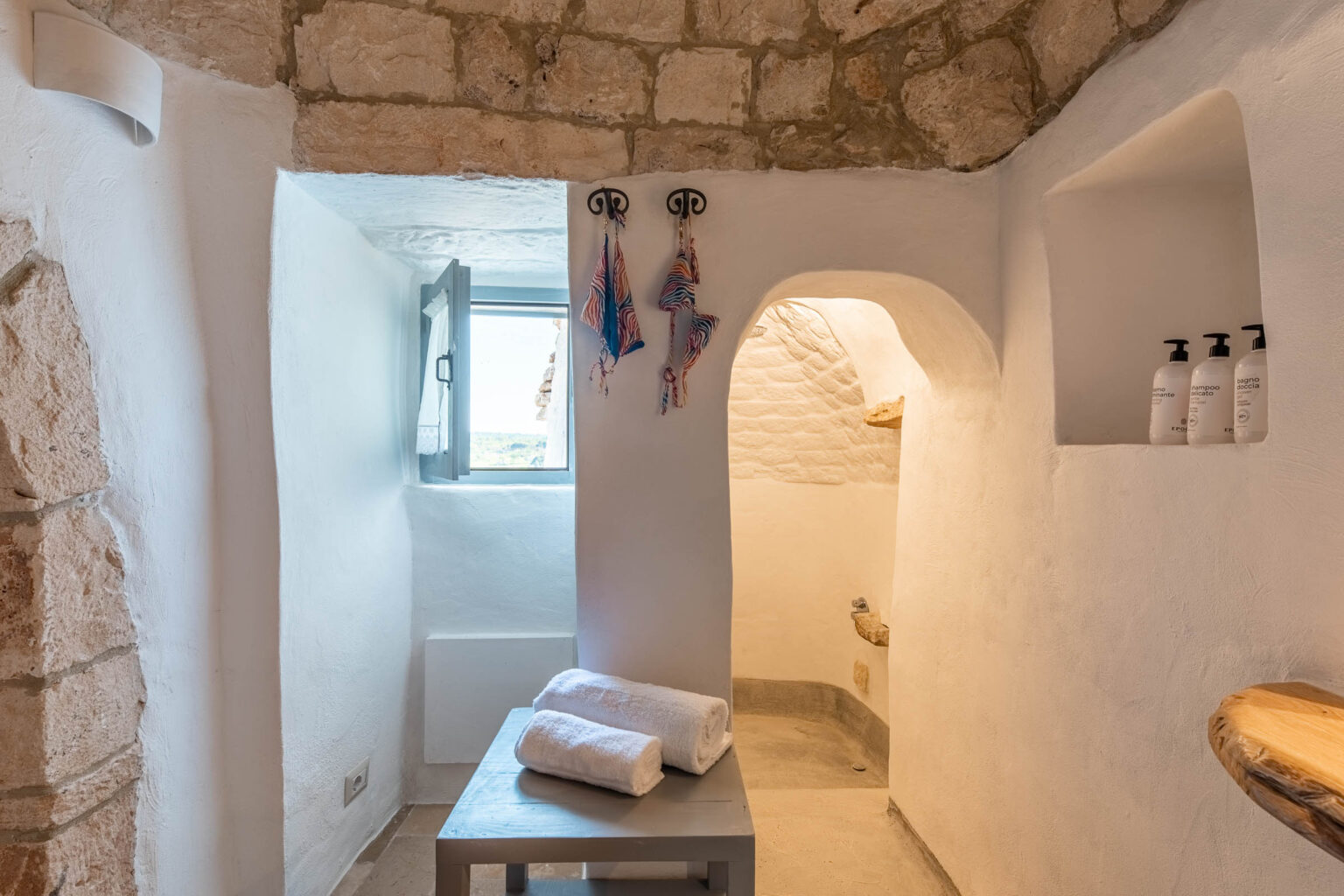 Interior view of a modern bathroom featuring stone walls, a small window, neatly arranged towels on a wooden table, and shower amenities on a shelf. The design combines rustic and contemporary elements, creating a serene and inviting atmosphere. | Only on SopranoVillas