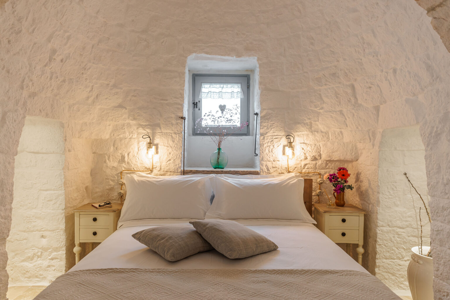 Cozy bedroom interior featuring a white stone wall, a comfortable double bed with plush pillows, and bedside tables with decorative flowers. Soft lighting enhances the serene atmosphere, complemented by a small window allowing natural light to filter in. Ideal for relaxation and a peaceful retreat. | Only on SopranoVillas
