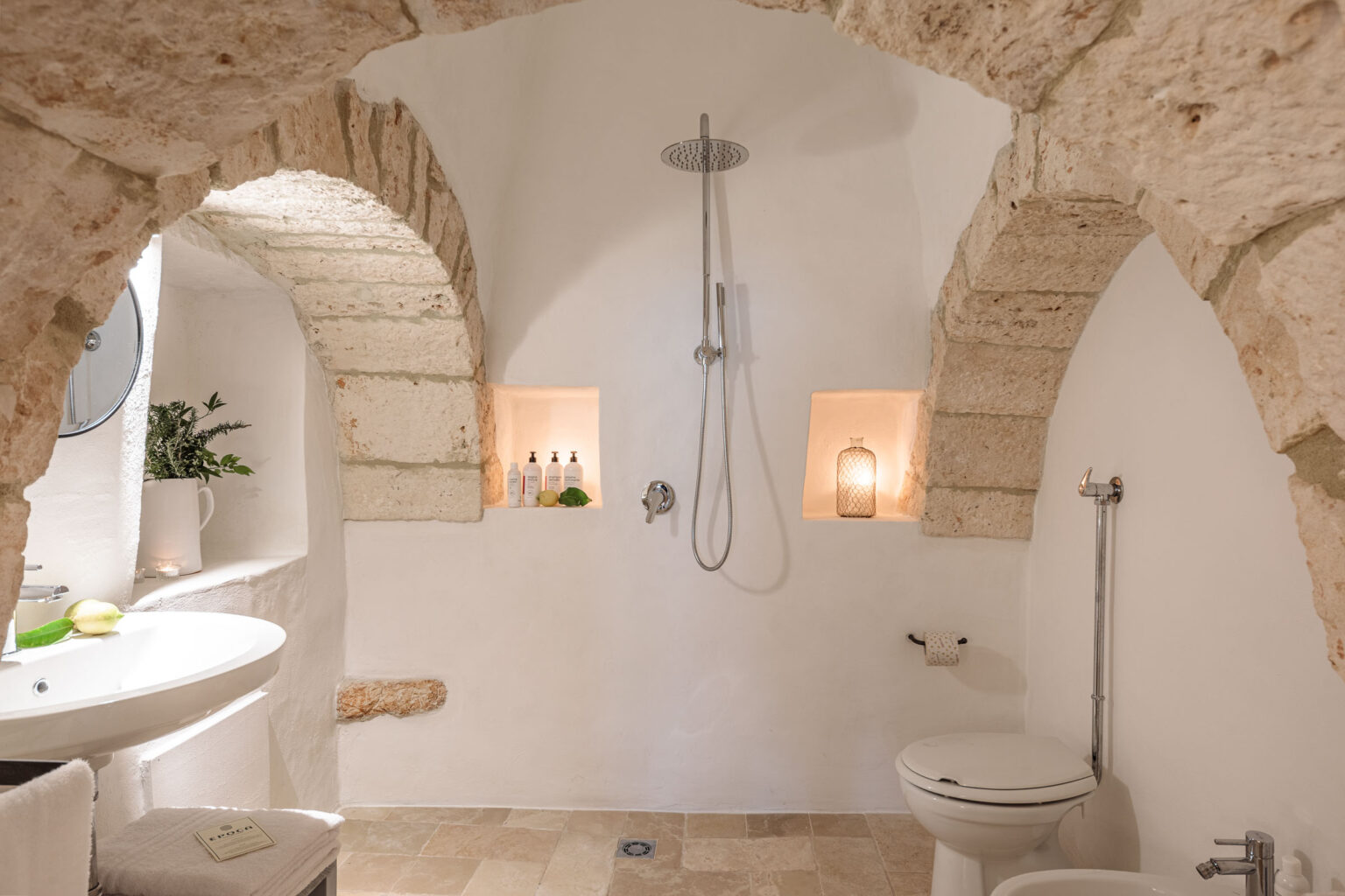 Modern bathroom design featuring arched stone architecture, a rainfall showerhead, and minimalist decor. The space includes a round mirror, a stylish sink, and illuminated shelves showcasing bath products and decorative elements, all set against a neutral color palette. | Only on SopranoVillas