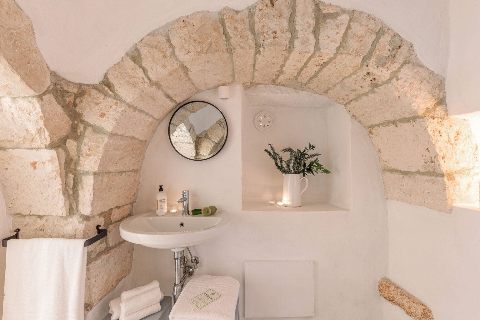 Elegant bathroom featuring a stone archway, round mirror, modern sink, and neatly arranged towels. The decor includes a white pitcher with greenery and candles, creating a serene and inviting atmosphere. Ideal for showcasing rustic charm in home design. | Only on SopranoVillas