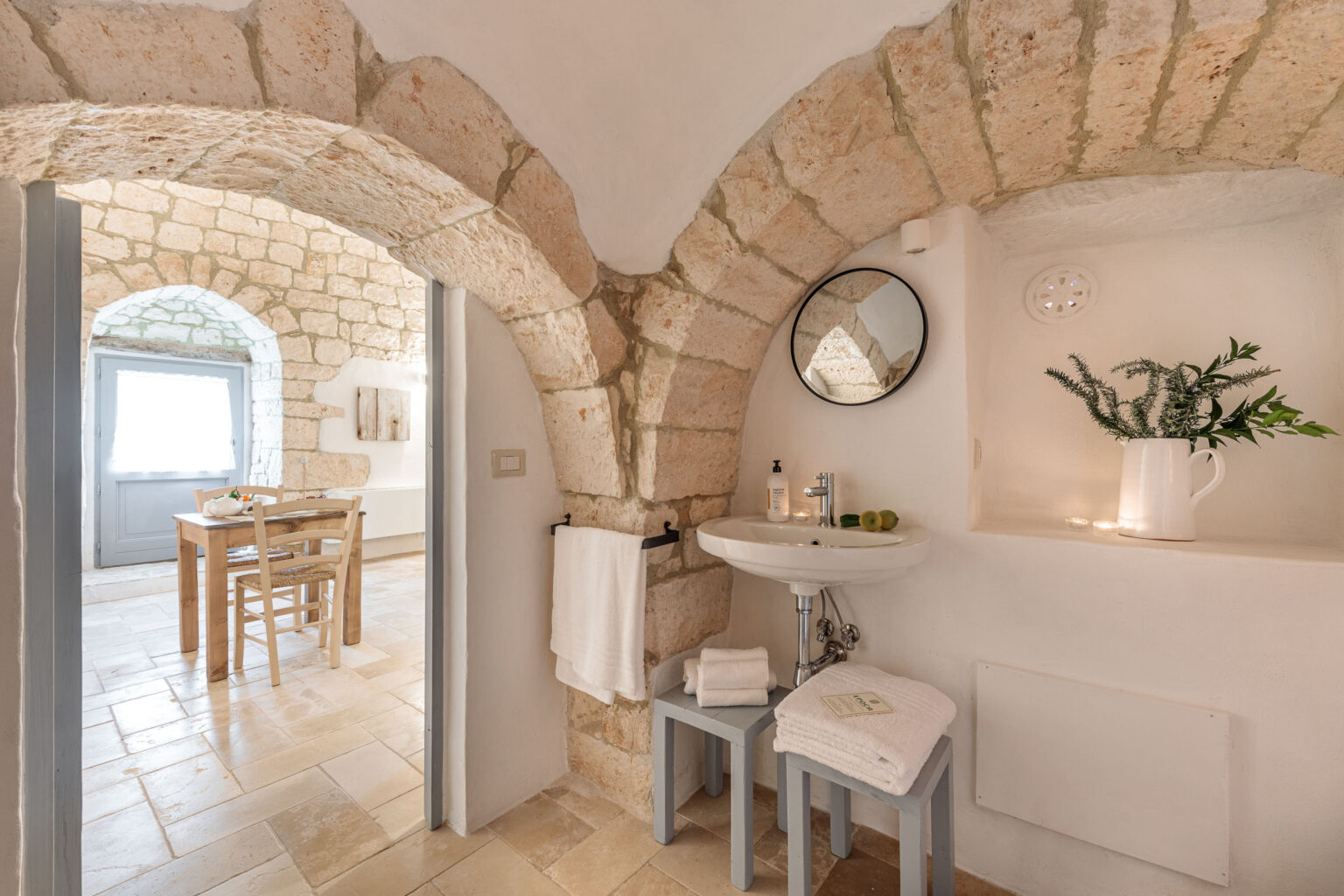 Cozy bathroom with stone archways featuring a round mirror, a white sink, and neatly arranged towels. Natural light illuminates the space, highlighting the rustic decor, including a small vase with greenery and a bowl of green apples. | Only on SopranoVillas