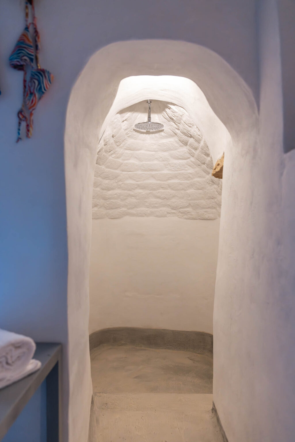 A serene shower area featuring an arched entrance, whitewashed walls, and a modern overhead showerhead, designed for a minimalist and relaxing atmosphere. Soft lighting enhances the tranquil vibe, ideal for a spa-like experience. | Only on SopranoVillas
