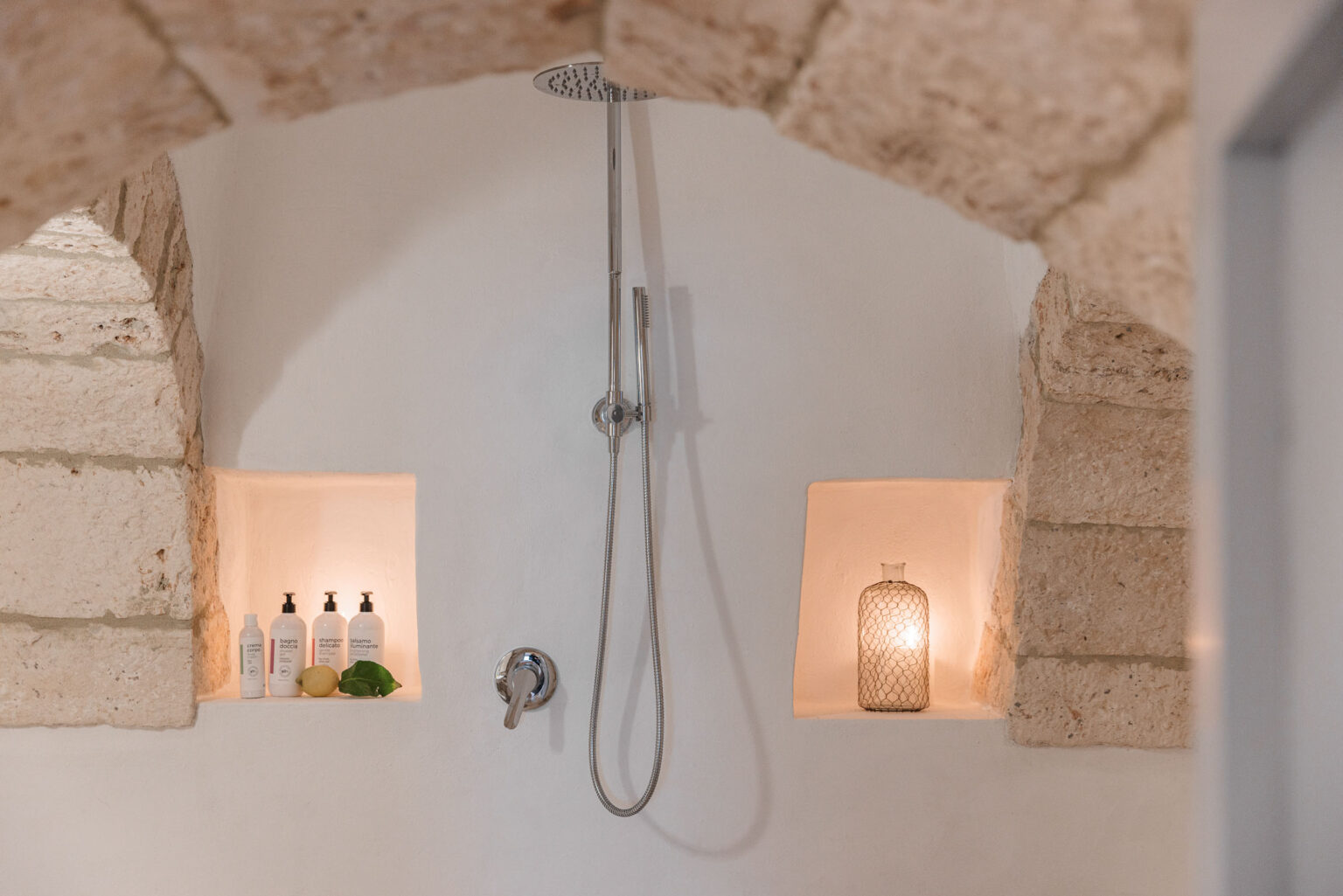 Modern shower area featuring a sleek overhead showerhead, illuminated alcoves with skincare products, and decorative candle holder, all set against a minimalist white wall and natural stone accents. | Only on SopranoVillas