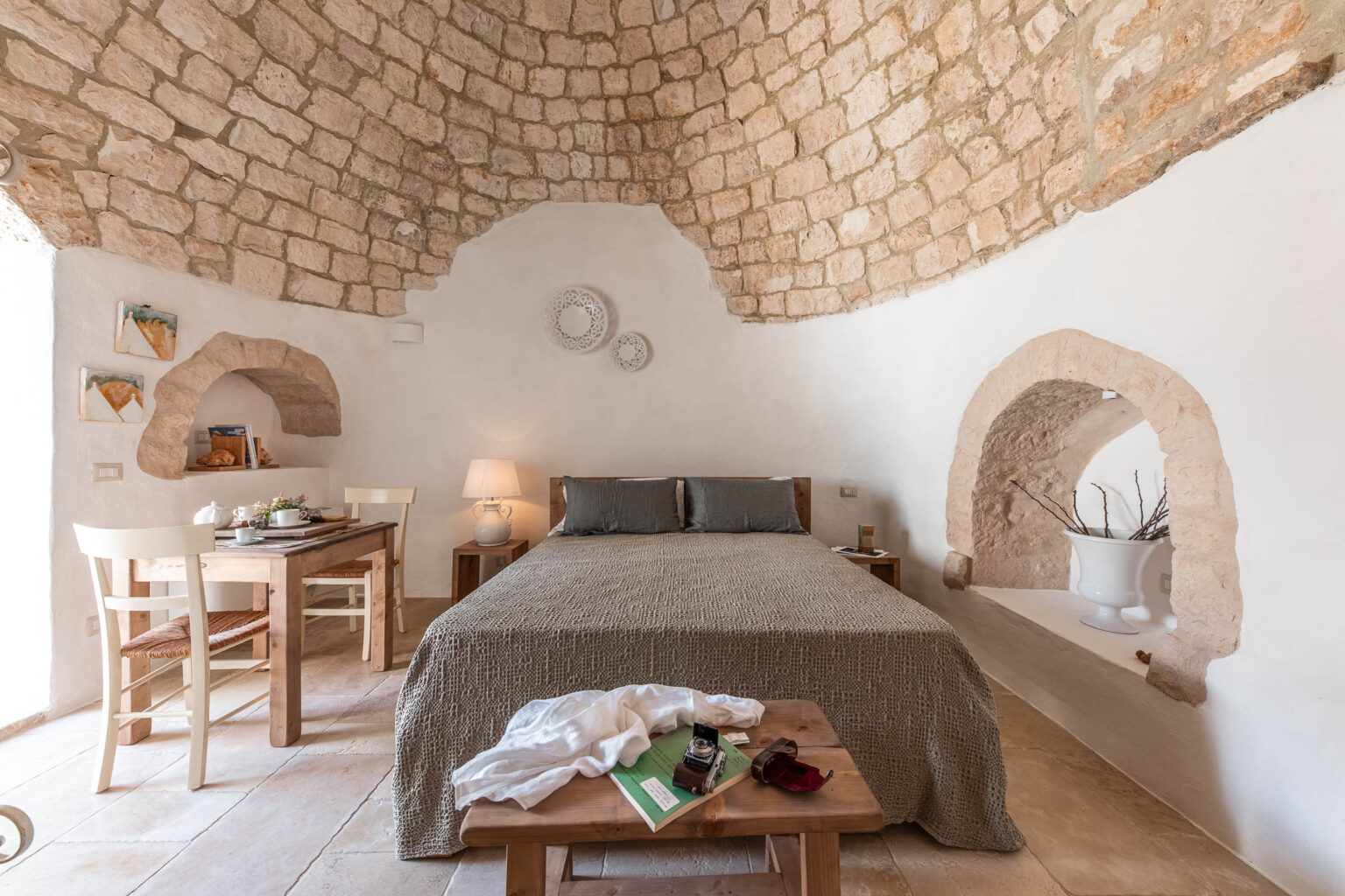 Cozy bedroom interior featuring a stone arched ceiling, a wooden bed with gray bedding, a rustic table with chairs, and decorative elements like potted plants and artwork on the walls. The room's neutral color palette and natural materials create a warm and inviting atmosphere. | Only on SopranoVillas