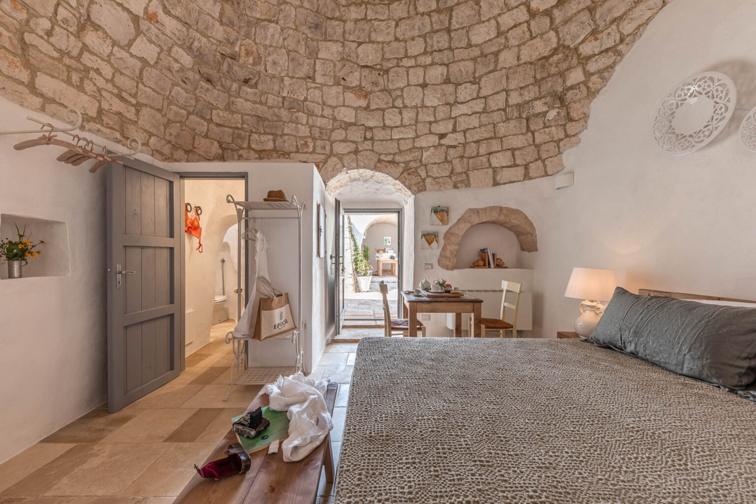 Cozy bedroom in a stone-walled cottage featuring a textured bedspread, wooden furniture, and a warm ambiance. The room includes a dining area, an arched doorway leading outside, and a view of a charming outdoor space. | Only on SopranoVillas