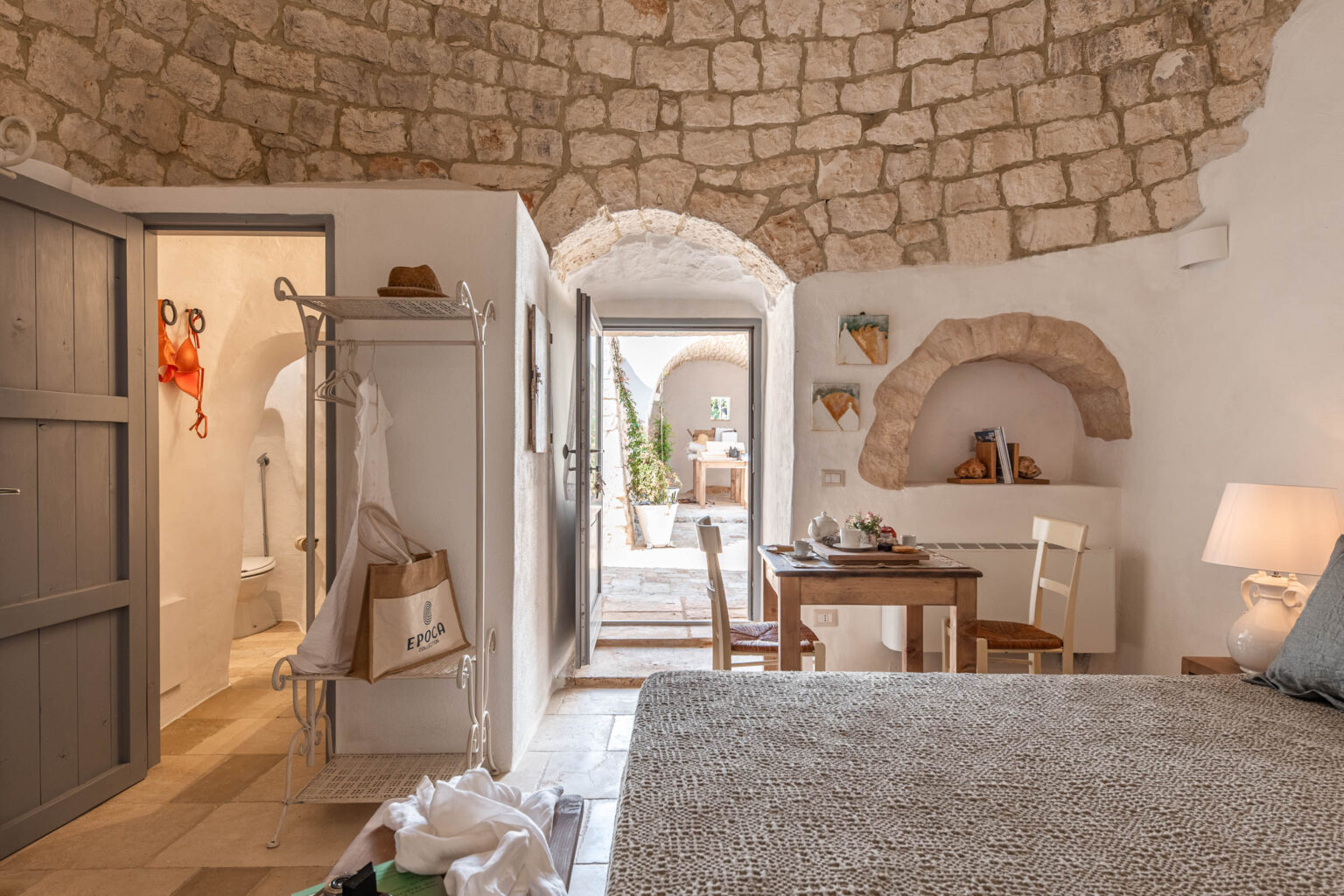 Cozy interior of a rustic bedroom featuring stone walls, a wooden table set for breakfast, and a view of an outdoor patio. The room includes a wardrobe with clothing, a comfortable bed with textured bedding, and decorative elements adding charm to the space. | Only on SopranoVillas