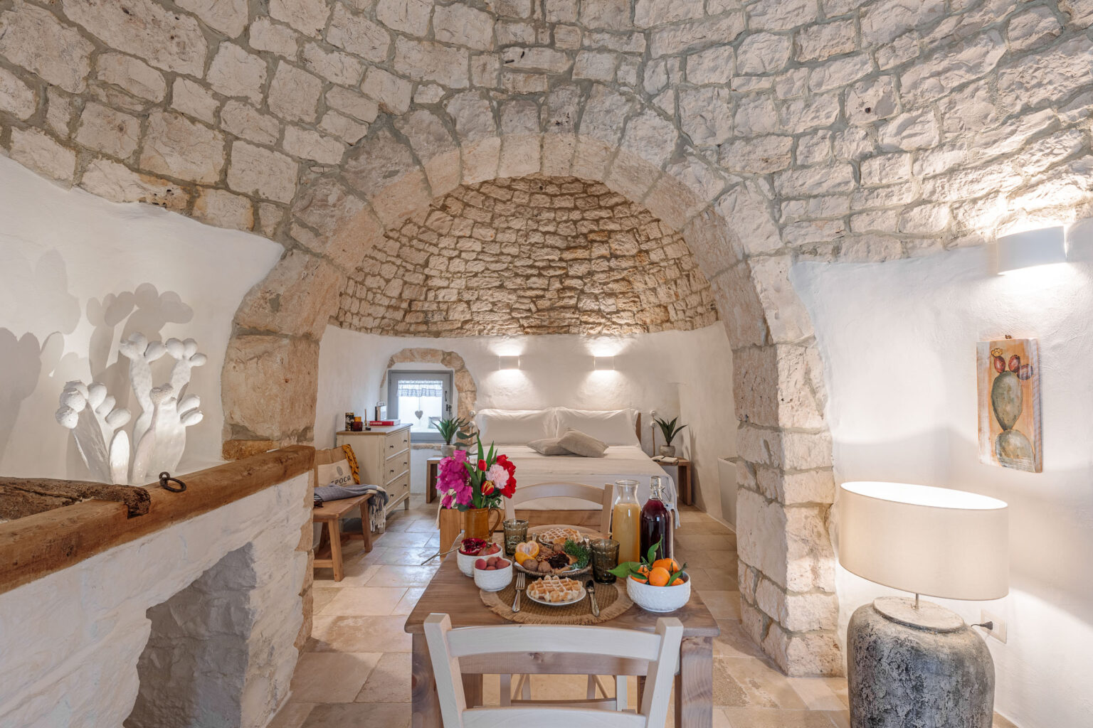 Cozy interior of a rustic stone cottage featuring a vaulted ceiling, a dining table set with breakfast items, and a comfortable bedroom area. The decor includes potted plants, a modern lamp, and colorful flowers, creating a warm and inviting atmosphere. | Only on SopranoVillas