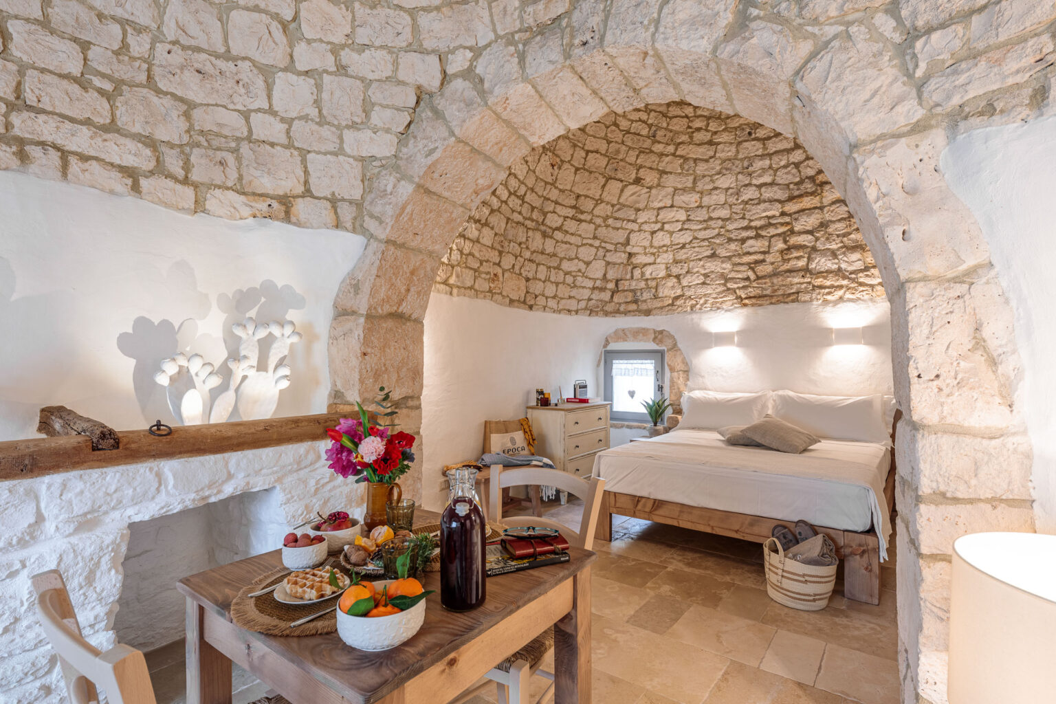 Cozy interior of a rustic bedroom featuring stone walls and arched ceilings, with a comfortable bed, a wooden dining table set with fresh fruits and pastries, and vibrant flowers creating a warm atmosphere. Natural light illuminates the space, enhancing its inviting charm. | Only on SopranoVillas
