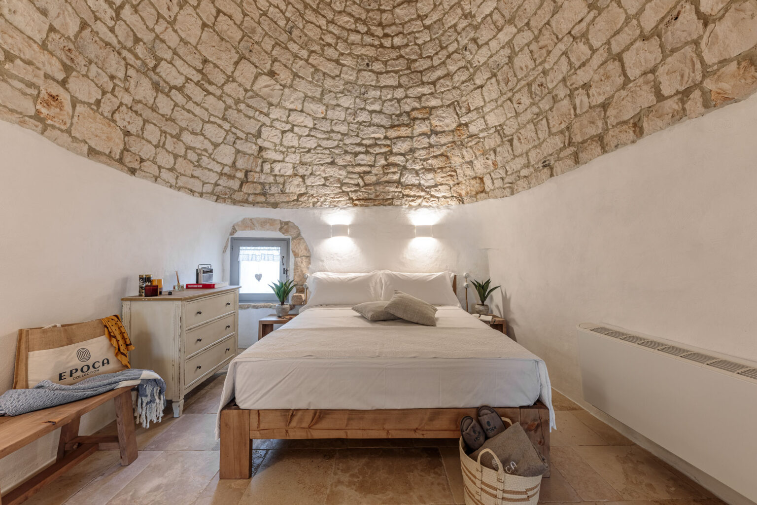 Cozy bedroom featuring a stone arch ceiling, a comfortable wooden bed with white linens, and minimalistic decor. The room includes a wooden dresser, a small window with a heart-shaped design, and potted plants for a touch of greenery. Natural light enhances the serene atmosphere, making it an inviting space for relaxation. | Only on SopranoVillas