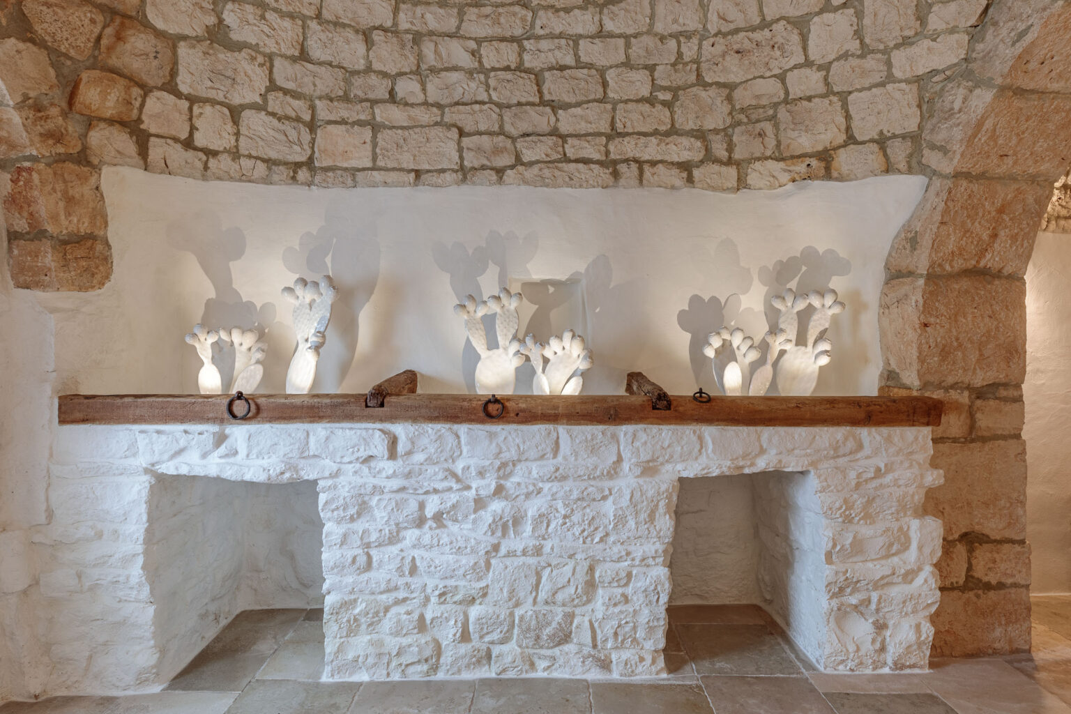 Interior view of a rustic stone wall with a wooden shelf displaying decorative white sculptures, illuminated to cast soft shadows, creating a warm and inviting atmosphere. The scene highlights the blend of natural materials and artistic design in a cozy setting. | Only on SopranoVillas