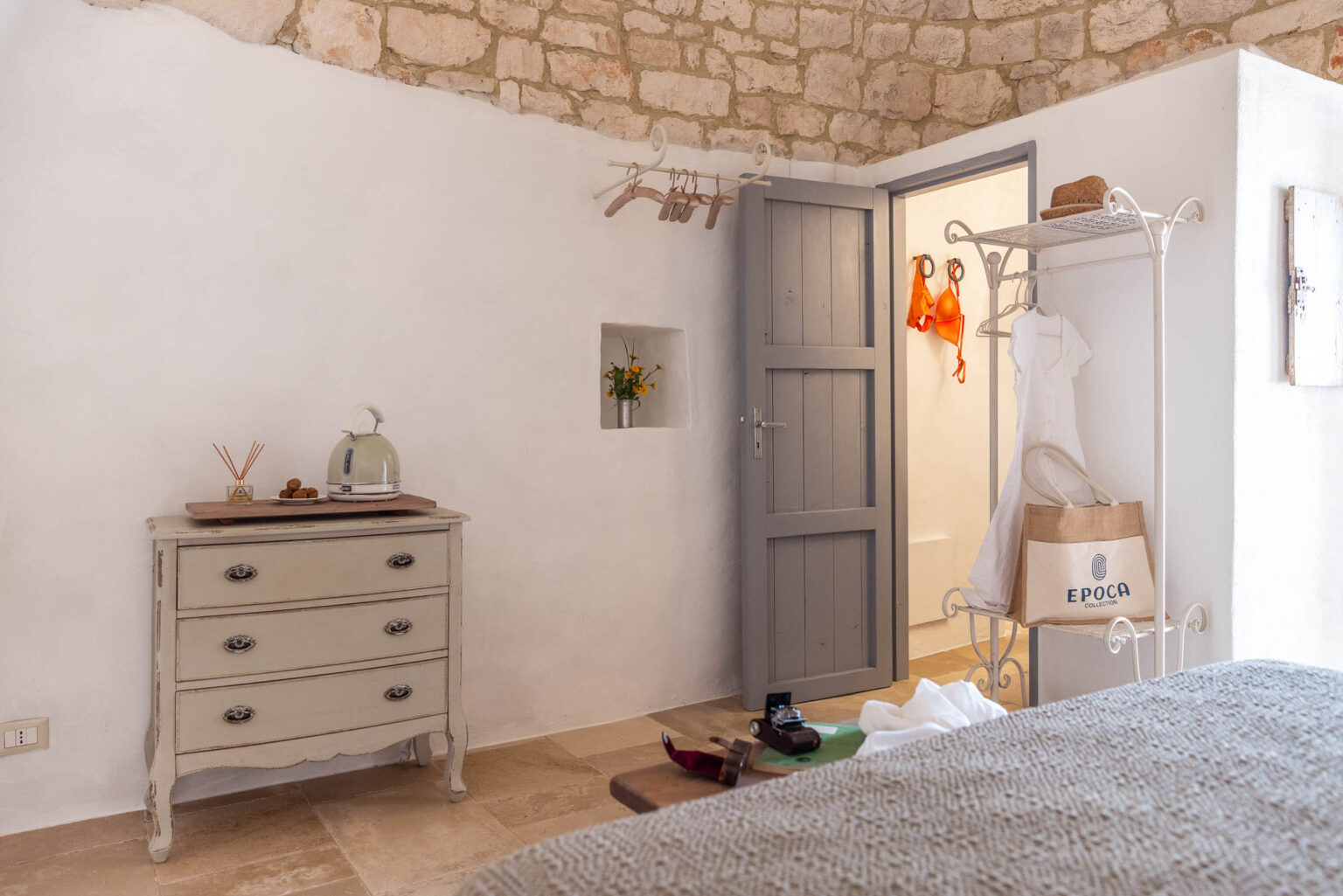 Cozy interior of a stylish bedroom featuring a vintage dresser, a kettle, and a decorative flower vase. A light dress hangs on a rack next to a door leading to a bathroom. The room has a warm, inviting atmosphere with stone walls and natural lighting. | Only on SopranoVillas
