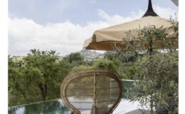 A stylish outdoor patio featuring a rattan chair, a modern teal side table, and a large beige umbrella, overlooking a serene infinity pool and lush greenery under a partly cloudy sky.