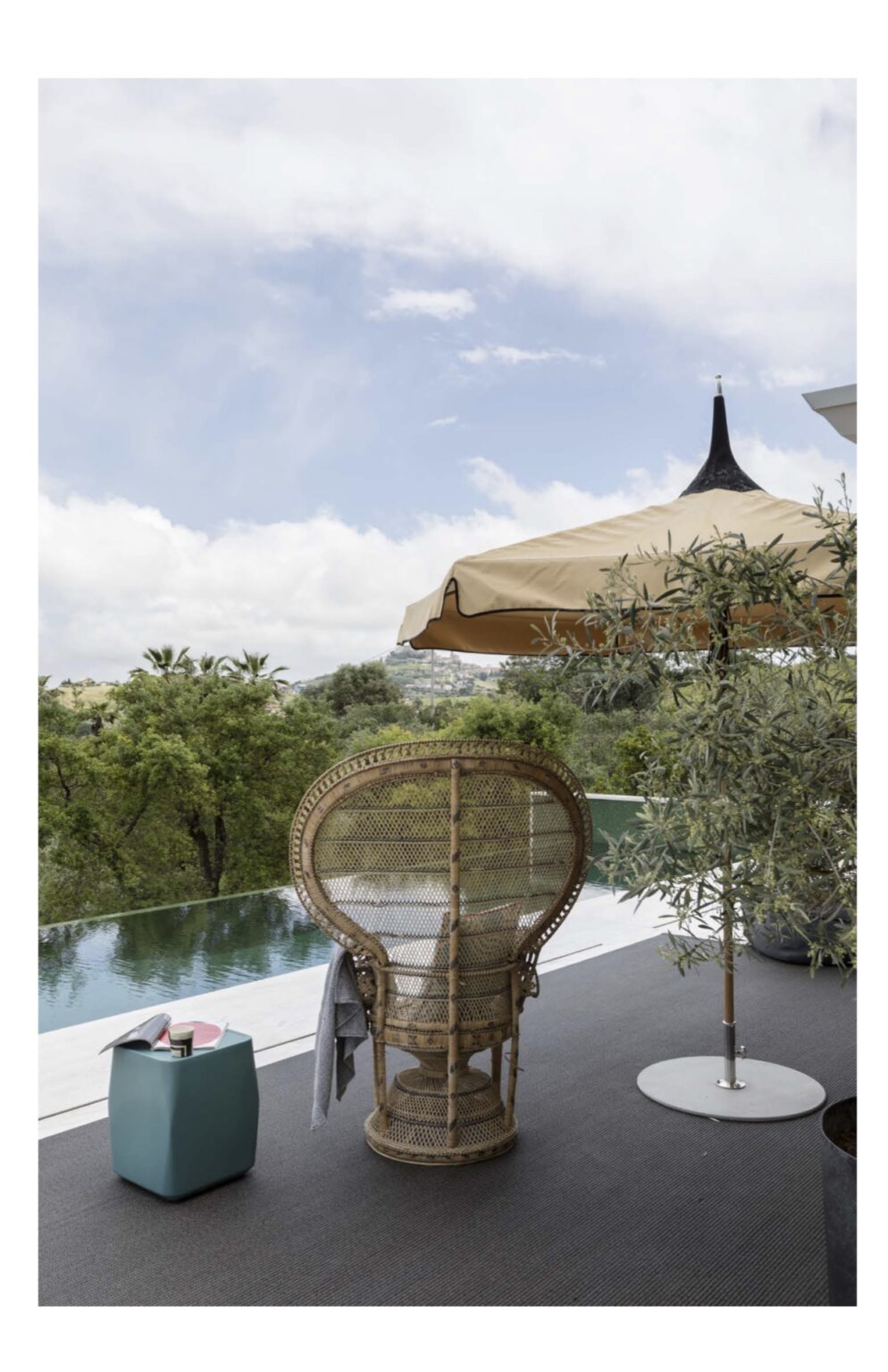 A stylish outdoor patio featuring a rattan chair, a modern teal side table, and a large beige umbrella, overlooking a serene infinity pool and lush greenery under a partly cloudy sky. | Only on SopranoVillas