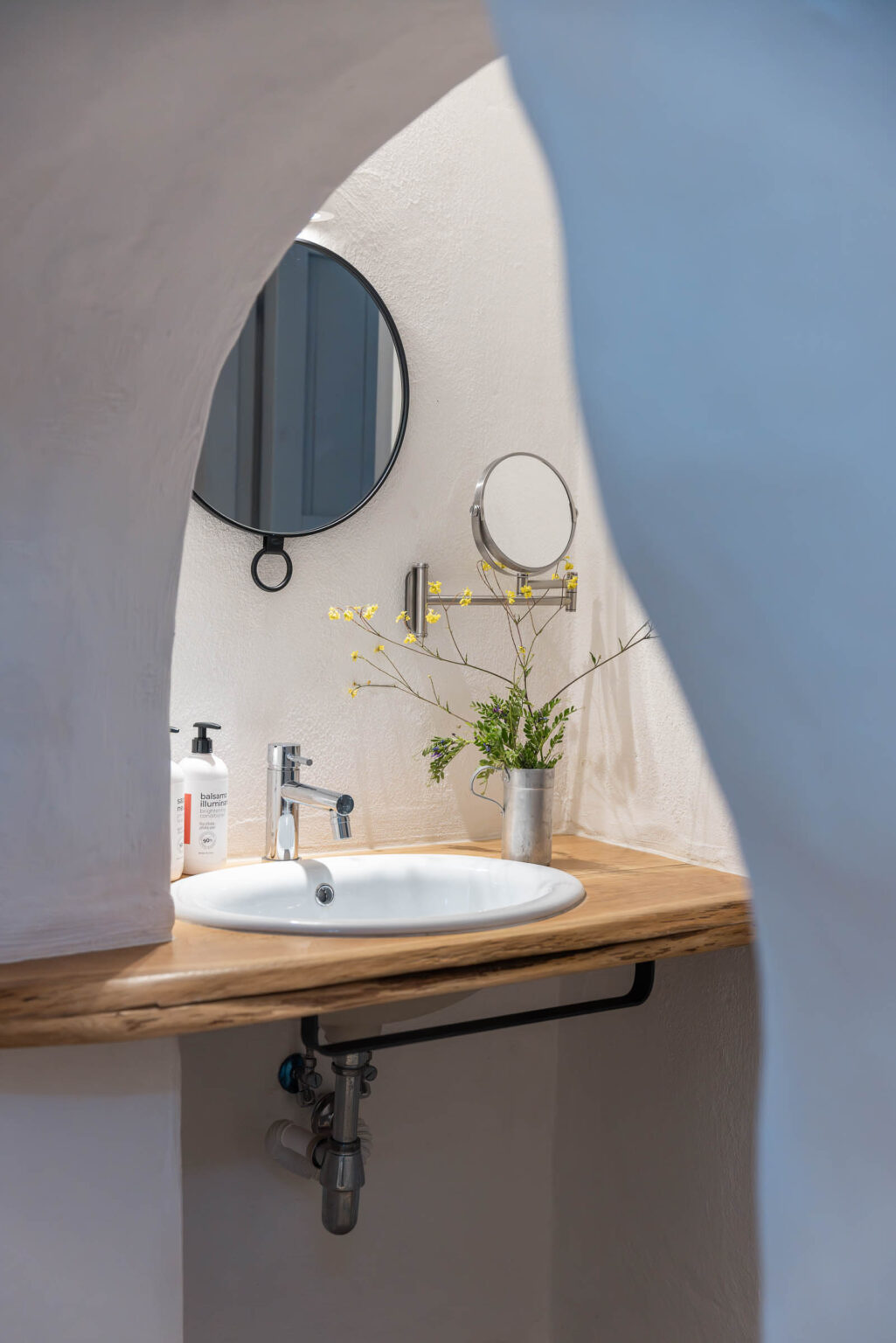 Modern bathroom interior featuring a sleek circular mirror, a stylish sink with a wooden countertop, and decorative flowers in a metal vase, creating a serene and inviting atmosphere. | Only on SopranoVillas