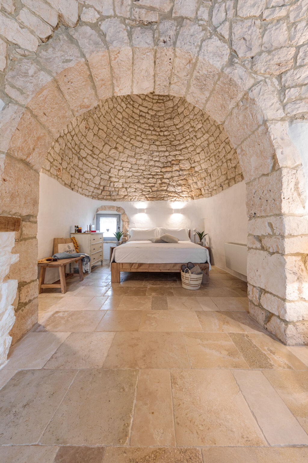 Interior view of a rustic bedroom featuring a stone arch ceiling, a wooden bed with white bedding, and minimalist decor. The room includes a dresser, a small table, and potted plants, set on beige stone flooring. Soft lighting enhances the cozy atmosphere. | Only on SopranoVillas