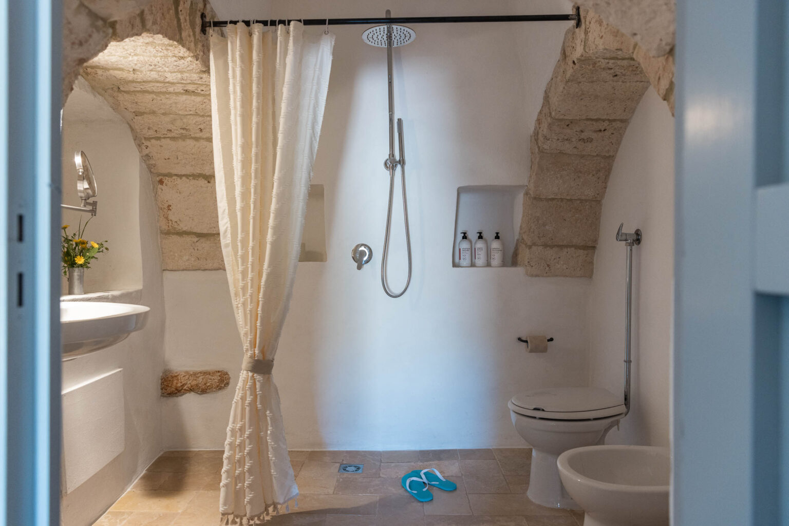 Modern bathroom featuring a shower with a rain head, a white sink, and a toilet. The space includes natural stone accents, a shower curtain, and toiletries displayed on a shelf. A pair of blue flip-flops is visible on the floor, adding a casual touch to the design. | Only on SopranoVillas