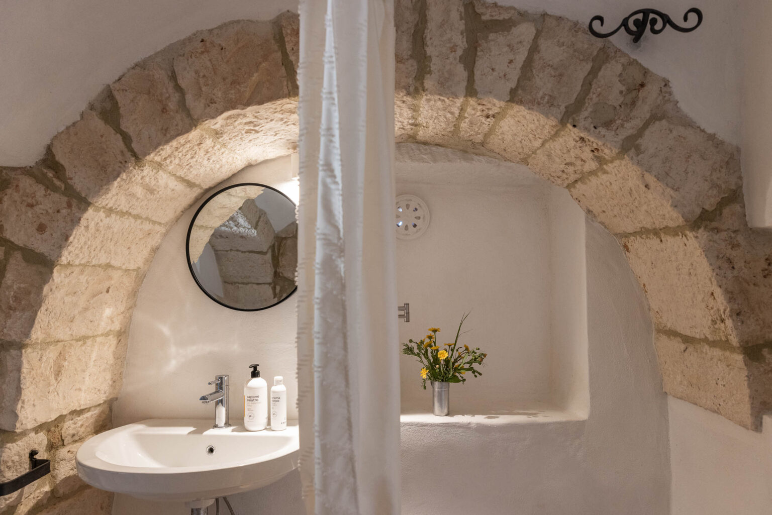 Modern bathroom featuring a stylish round mirror, a white sink with sleek faucets, and decorative stone archways. Two bottles of shampoo and conditioner are displayed on the counter, alongside a small vase of fresh flowers, creating a serene and inviting atmosphere. | Only on SopranoVillas