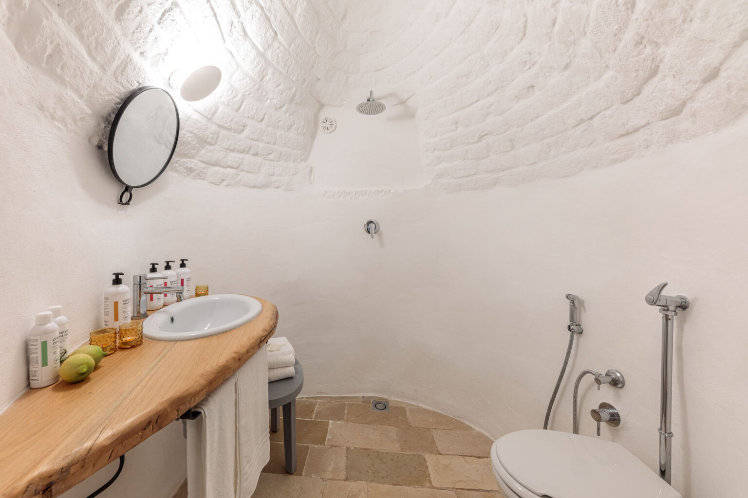 Modern bathroom interior featuring a curved stone ceiling, a wooden vanity with a round mirror, and neatly arranged toiletries. The space includes a shower, a toilet, and natural stone flooring, creating a minimalist and serene atmosphere. | Only on SopranoVillas
