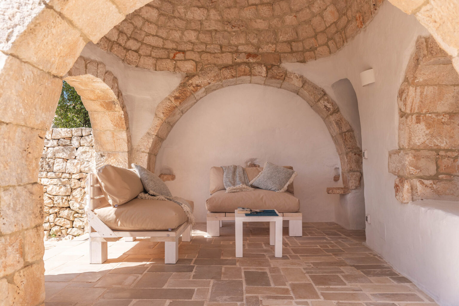 Cozy interior of a rustic stone room featuring arched ceilings, two beige lounge chairs with decorative pillows, and a small white table, illuminated by natural light filtering through a window. The floor is made of large stone tiles, enhancing the room's warm, inviting atmosphere. | Only on SopranoVillas