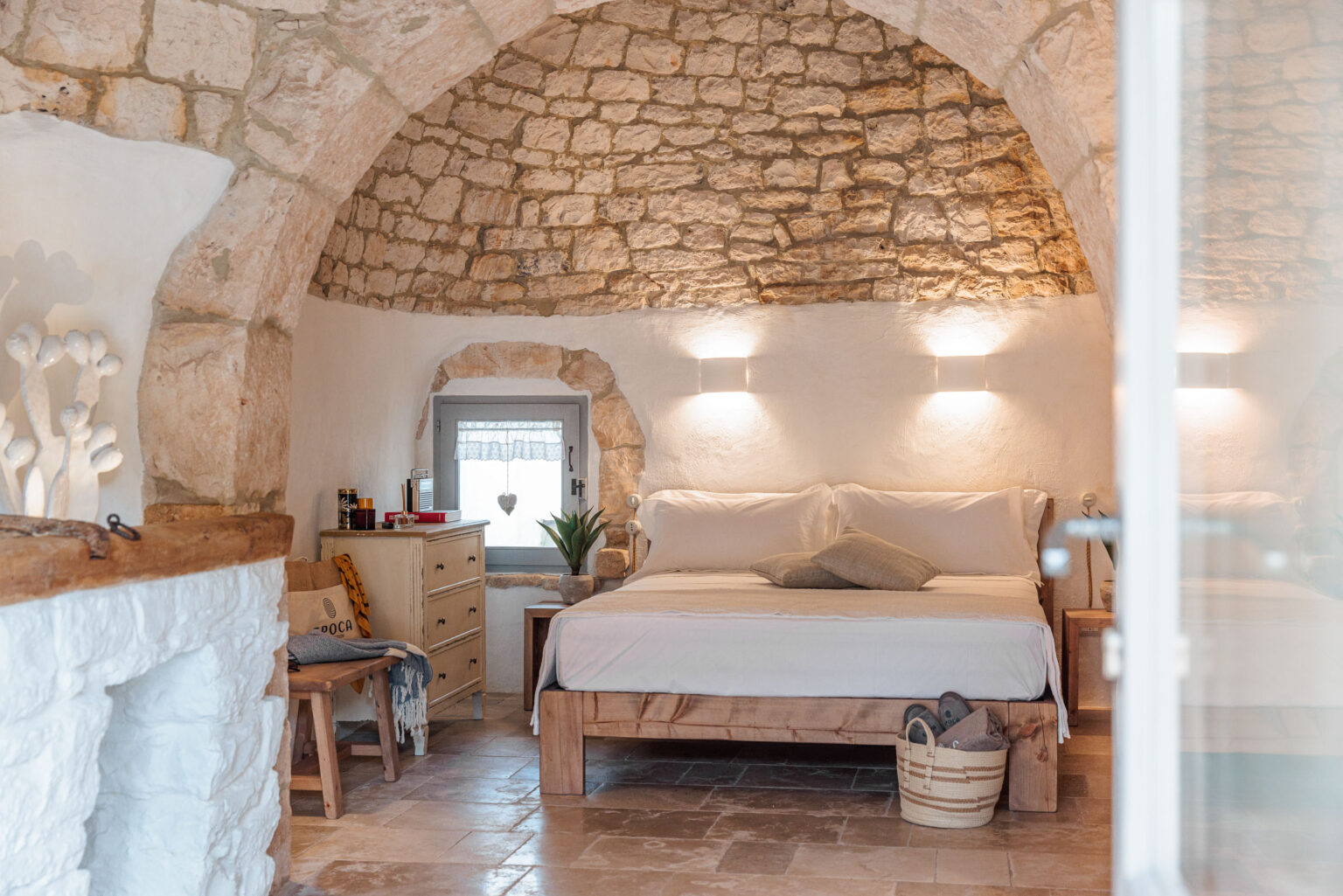 Cozy bedroom with arched stone ceiling, featuring a wooden bed with white bedding, bedside tables, and a decorative plant. A vintage dresser and a comfortable chair complete the rustic decor, while natural light enters through a small window adorned with a lace curtain. | Only on SopranoVillas
