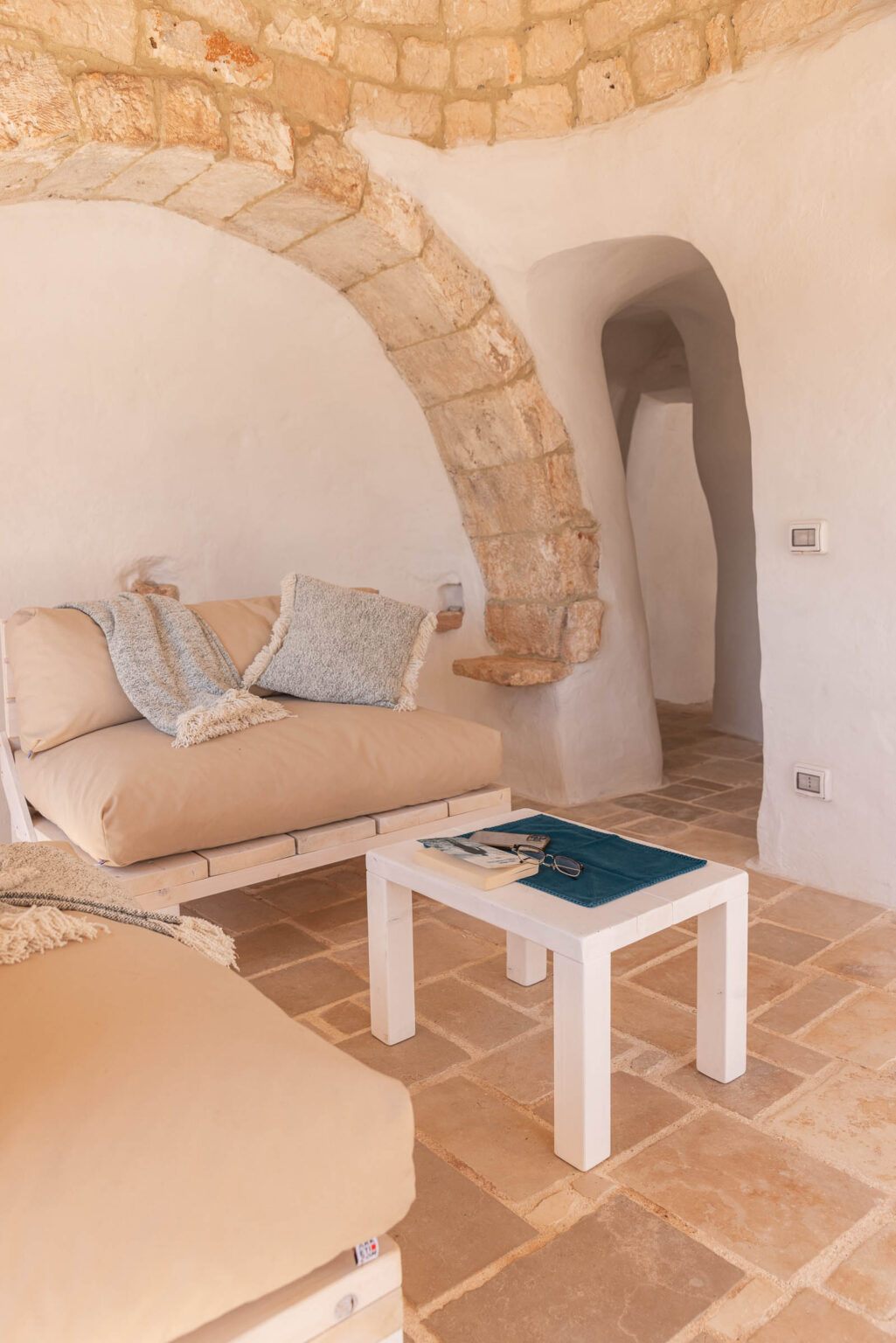 Cozy interior of a rustic living room featuring a beige couch with decorative pillows, a white coffee table with books and glasses, and a stone archway in a minimalist design. The floor is made of natural stone tiles, enhancing the warm and inviting atmosphere. | Only on SopranoVillas