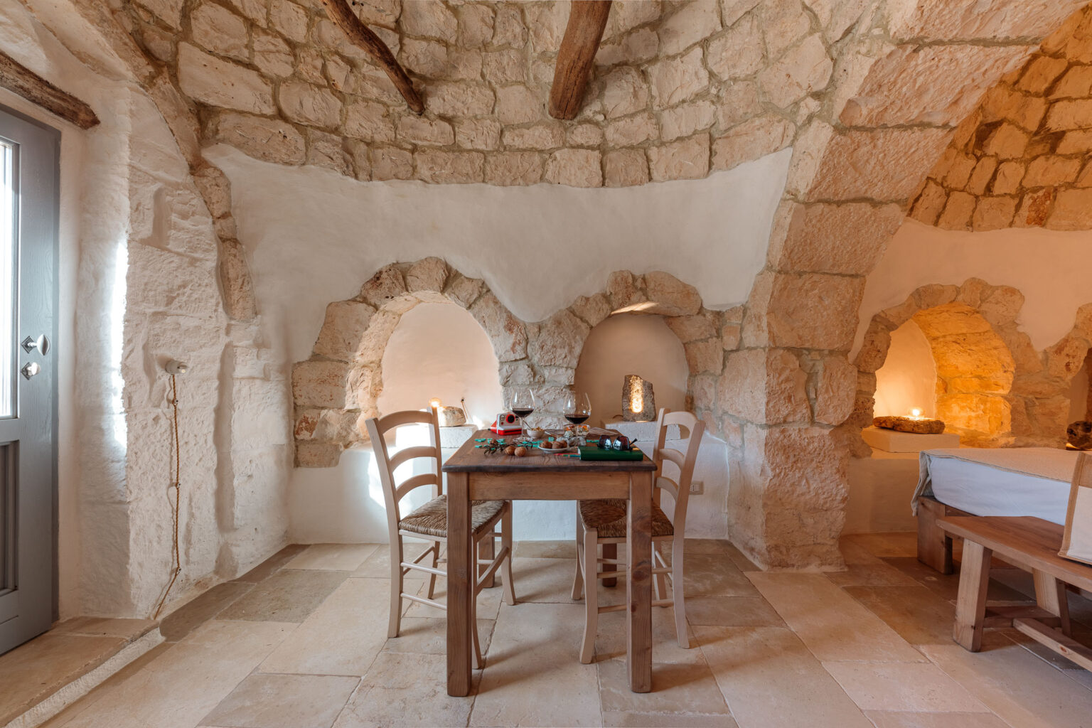 Cozy dining area featuring a rustic wooden table with two chairs, set against a backdrop of stone walls and arched niches. Natural light illuminates the space, enhancing the warm ambiance of the room. Ideal for intimate gatherings or a romantic dinner. | Only on SopranoVillas