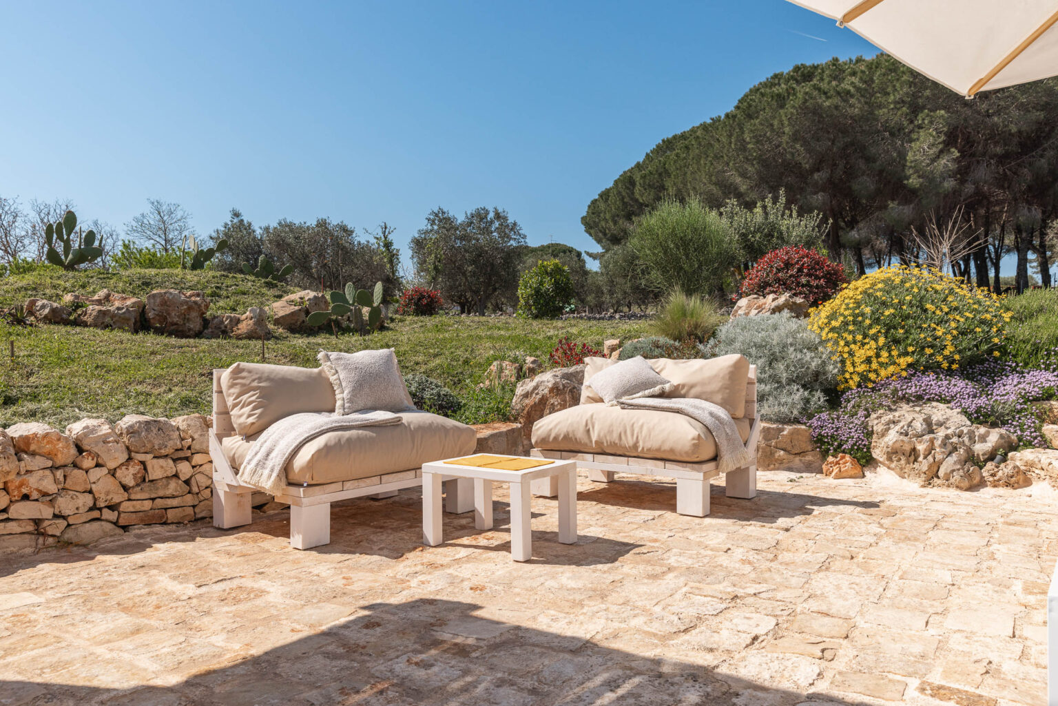 Cozy outdoor seating area with two beige cushioned chairs and a small white table, surrounded by lush greenery and colorful flowers under a clear blue sky. Ideal for relaxation and enjoying nature. | Only on SopranoVillas