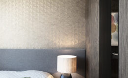 Modern bedroom interior featuring a textured wall, grey upholstered headboard, and a cozy bed with a green decorative pillow. A stylish lamp on a round side table adds warmth, while a partition hints at a bathroom space in the background.