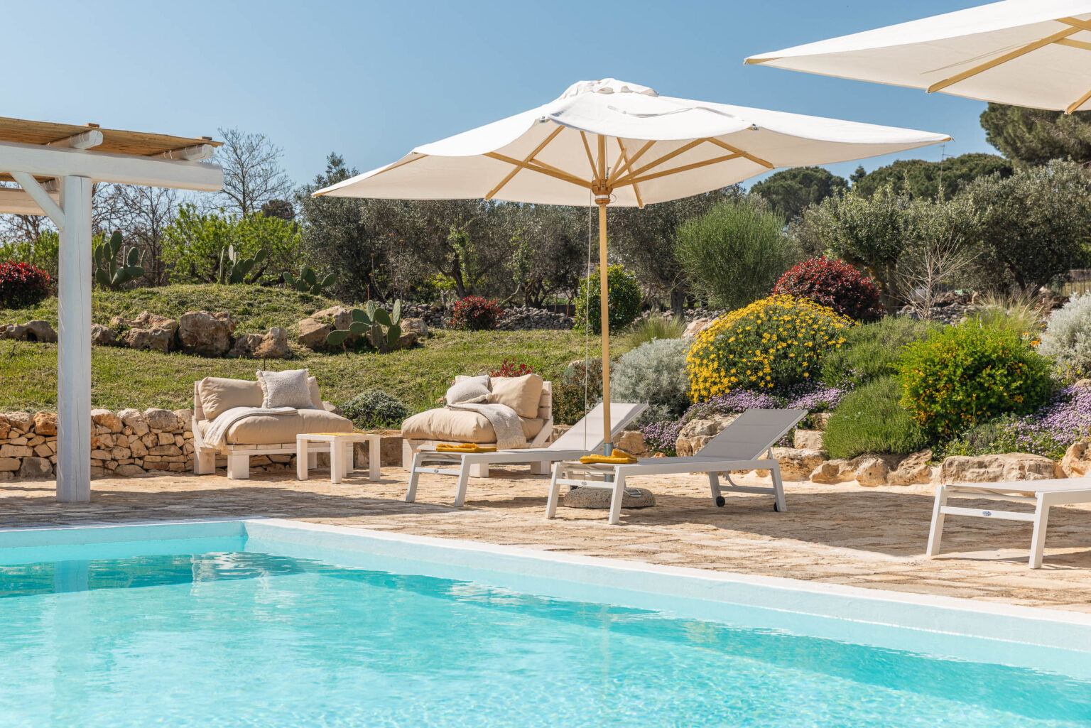 Luxury outdoor pool area featuring comfortable lounge chairs, large umbrellas, and vibrant flowers in a serene garden setting. Perfect for relaxation and leisure in sunny weather. | Only on SopranoVillas