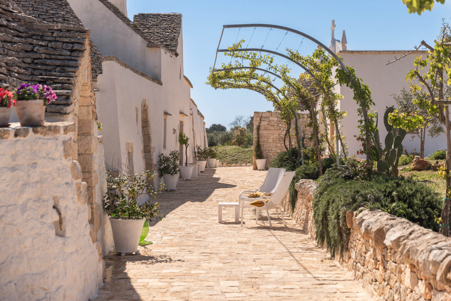 Charming stone pathway lined with potted plants and whitewashed buildings, featuring a modern seating area with chairs and a table, under a clear blue sky. | Only on SopranoVillas