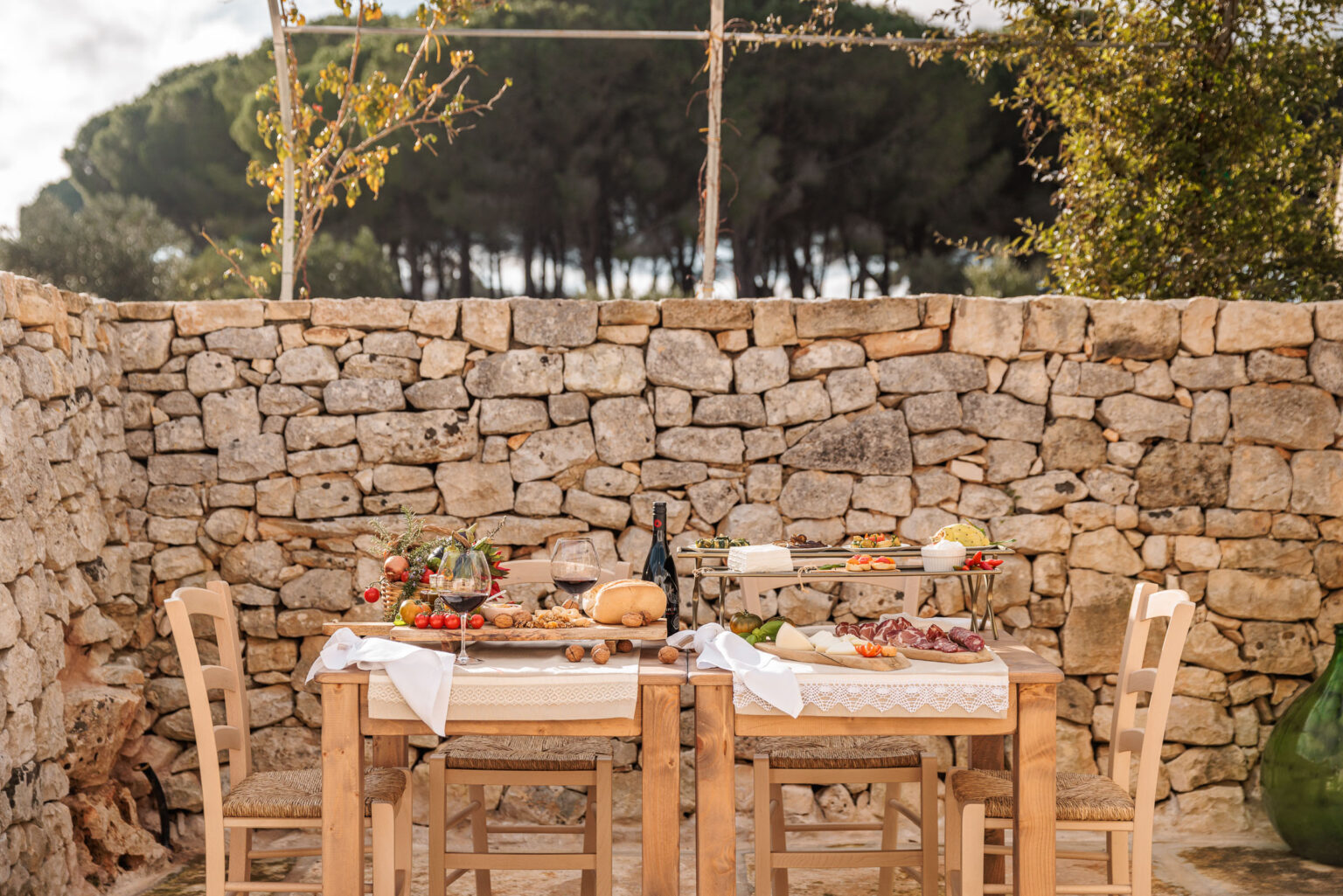 Outdoor dining setup featuring a rustic wooden table adorned with a variety of gourmet food, including cheese, meats, and fresh vegetables, complemented by wine glasses and a bottle of red wine. The scene is framed by a picturesque stone wall and lush greenery, creating an inviting atmosphere for a leisurely meal in nature. | Only on SopranoVillas