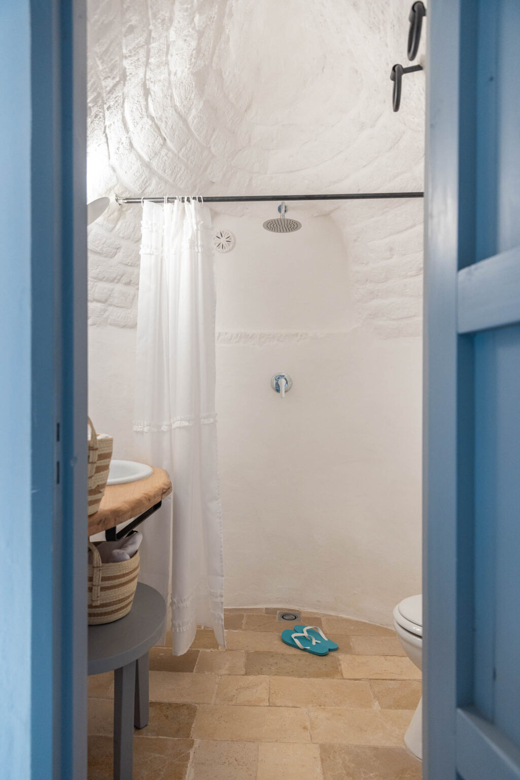 Bathroom interior featuring a unique curved ceiling, white walls, and natural stone flooring. A shower with a curtain, a circular sink on a wooden countertop, and a toilet are visible. Light blue door adds a pop of color, while teal flip-flops are placed on the floor. | Only on SopranoVillas