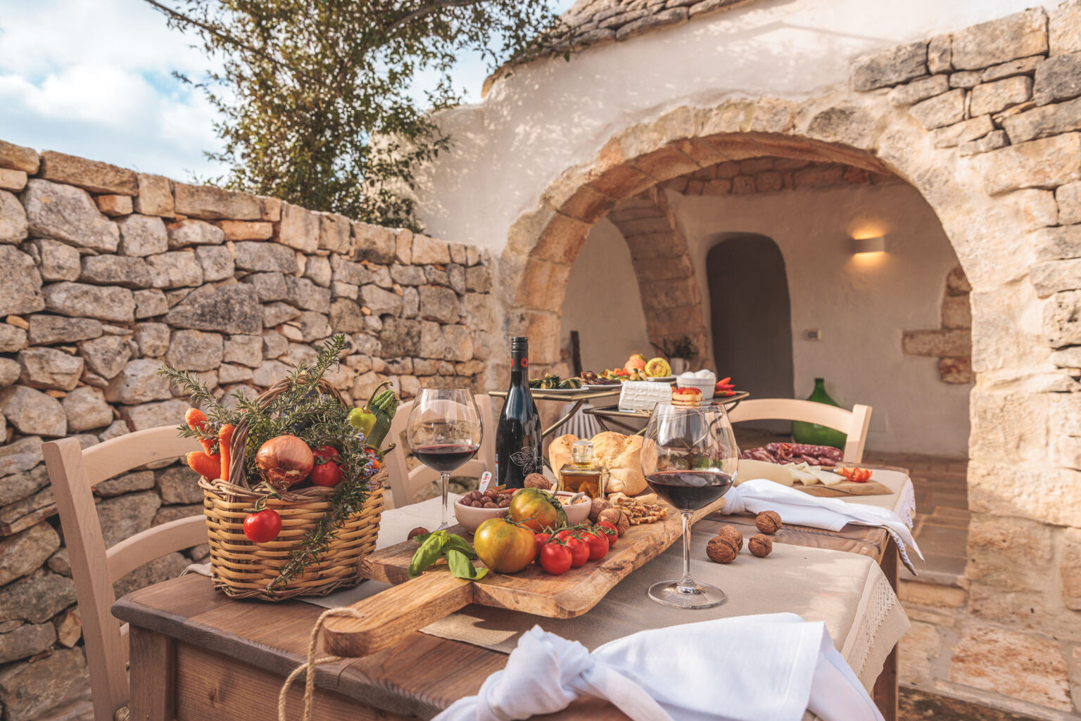 Rustic outdoor dining setup featuring a wooden table adorned with a basket of fresh vegetables, a cutting board with assorted tomatoes and bread, and glasses of red wine. Stone walls and an arched entrance create a charming backdrop for a Mediterranean-inspired meal. | Only on SopranoVillas
