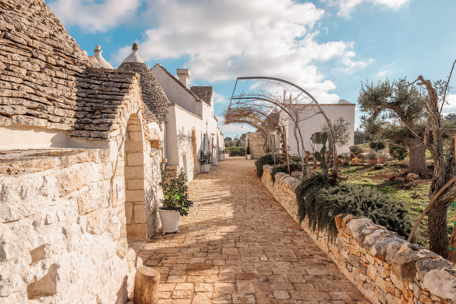 Charming stone pathway lined with traditional trulli houses in a picturesque Italian village, featuring lush greenery and a clear blue sky. | Only on SopranoVillas