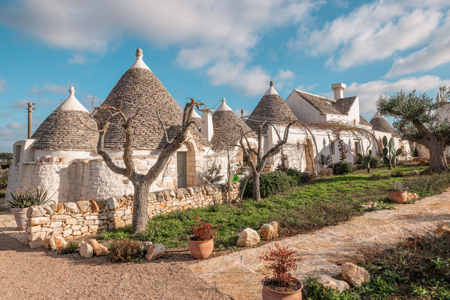 Trulli houses in Alberobello, Italy, showcasing traditional conical roofs and whitewashed stone walls, surrounded by greenery and a gravel pathway. | Only on SopranoVillas