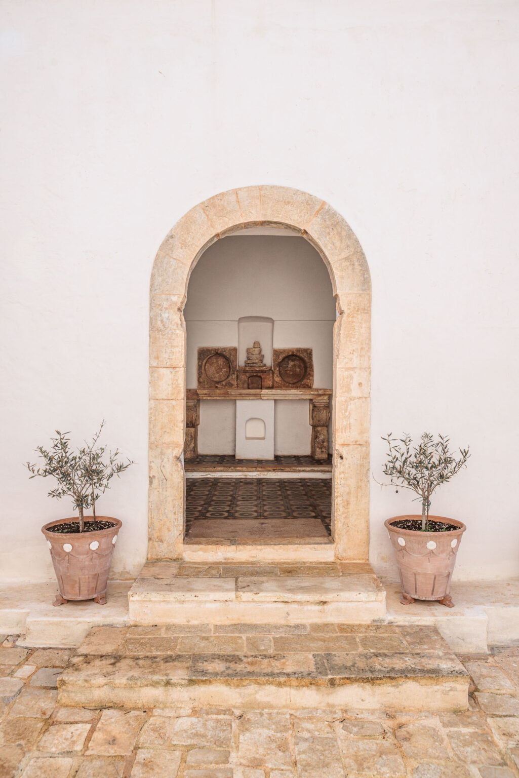 A stone archway leads to a minimalist interior with a decorative wall and traditional tiled floor. Two terracotta pots with olive trees flank the entrance, enhancing the serene ambiance of the setting. | Only on SopranoVillas