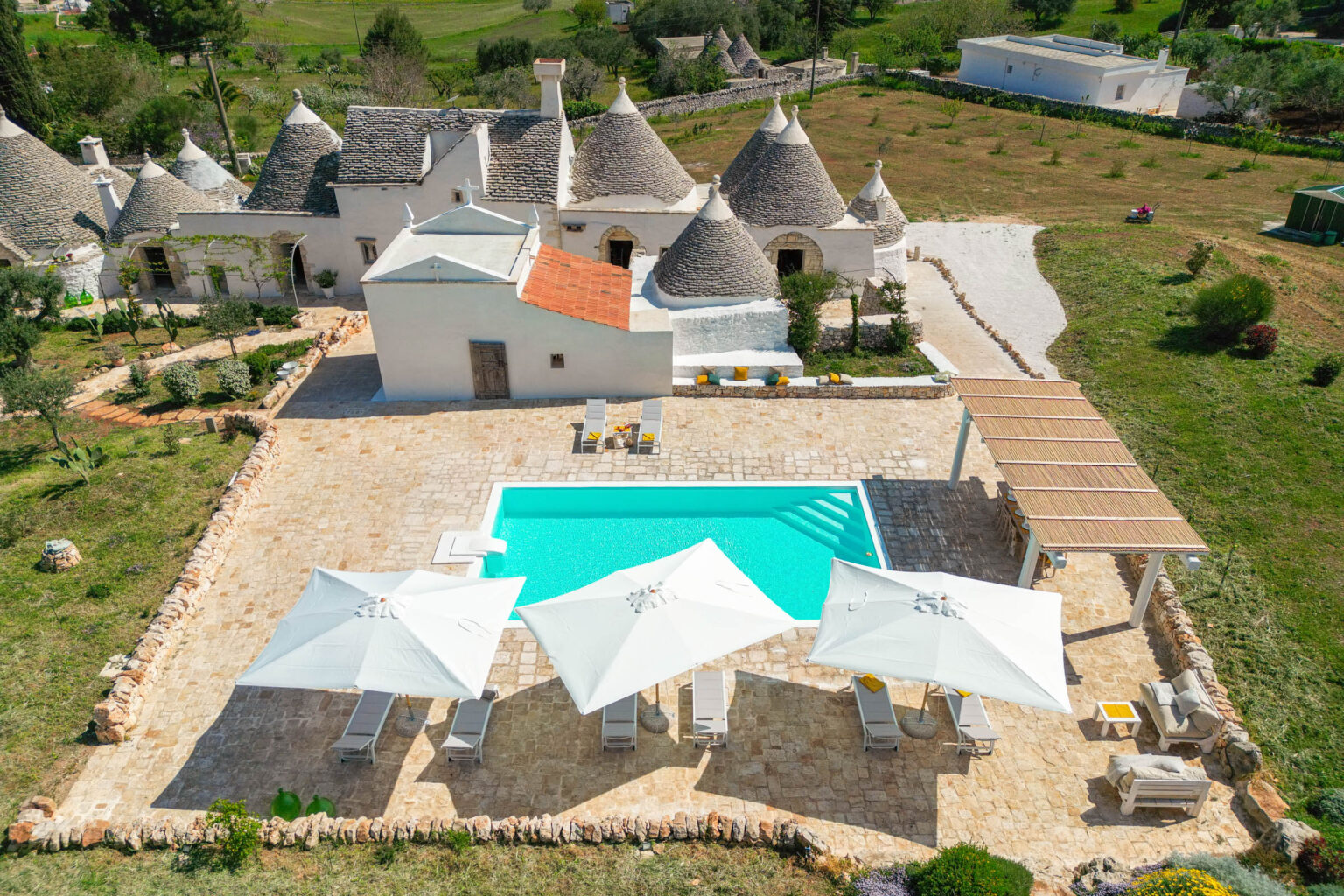 Aerial view of a traditional trullo house with conical roofs surrounded by lush greenery, featuring a swimming pool with lounge chairs and umbrellas in a spacious outdoor area. The property showcases a blend of natural beauty and rustic architecture, ideal for relaxation and leisure. | Only on SopranoVillas