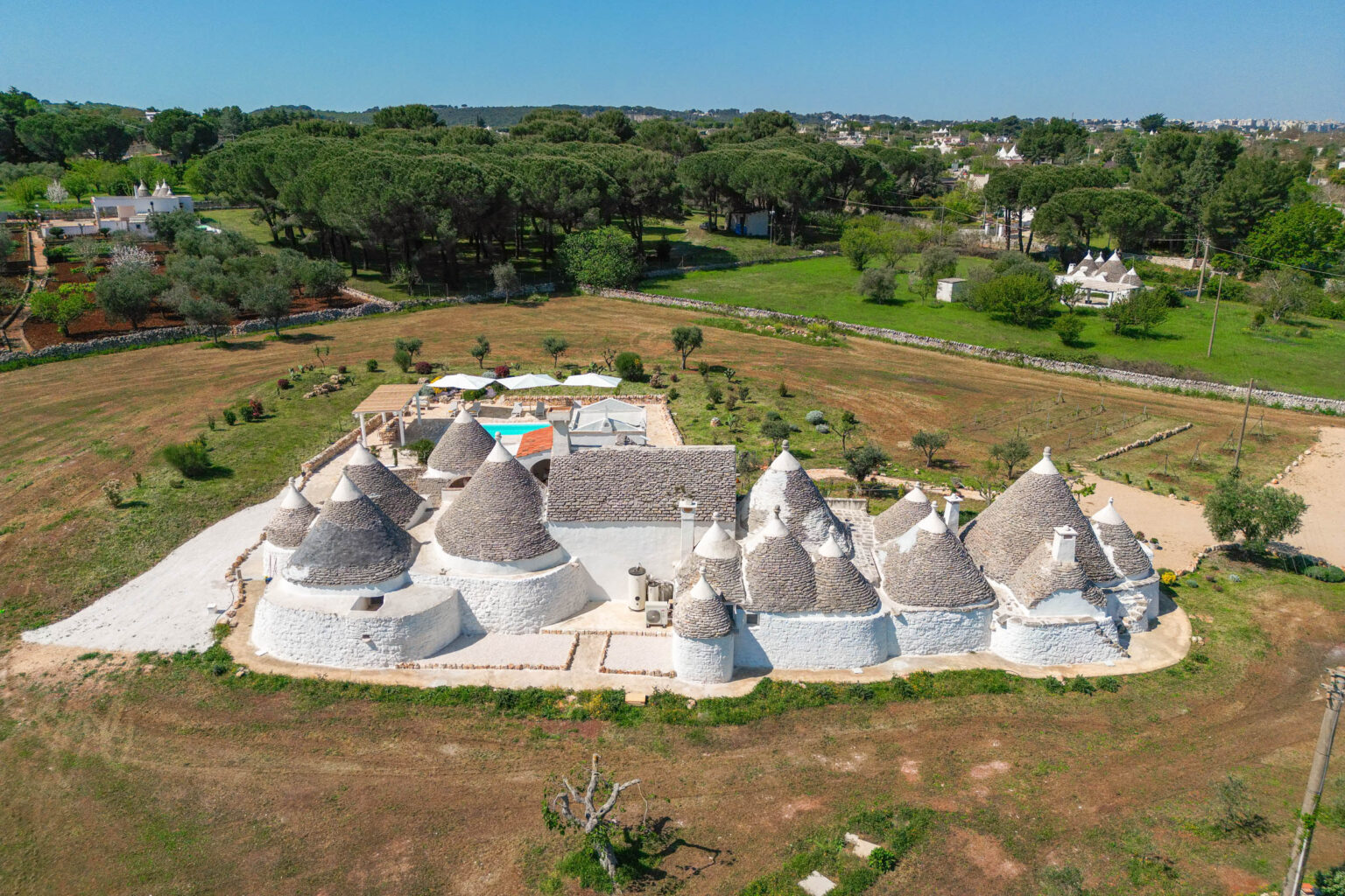 Aerial view of traditional trulli houses in Puglia, Italy, surrounded by lush greenery and gardens, showcasing their unique conical roofs and whitewashed walls. | Only on SopranoVillas