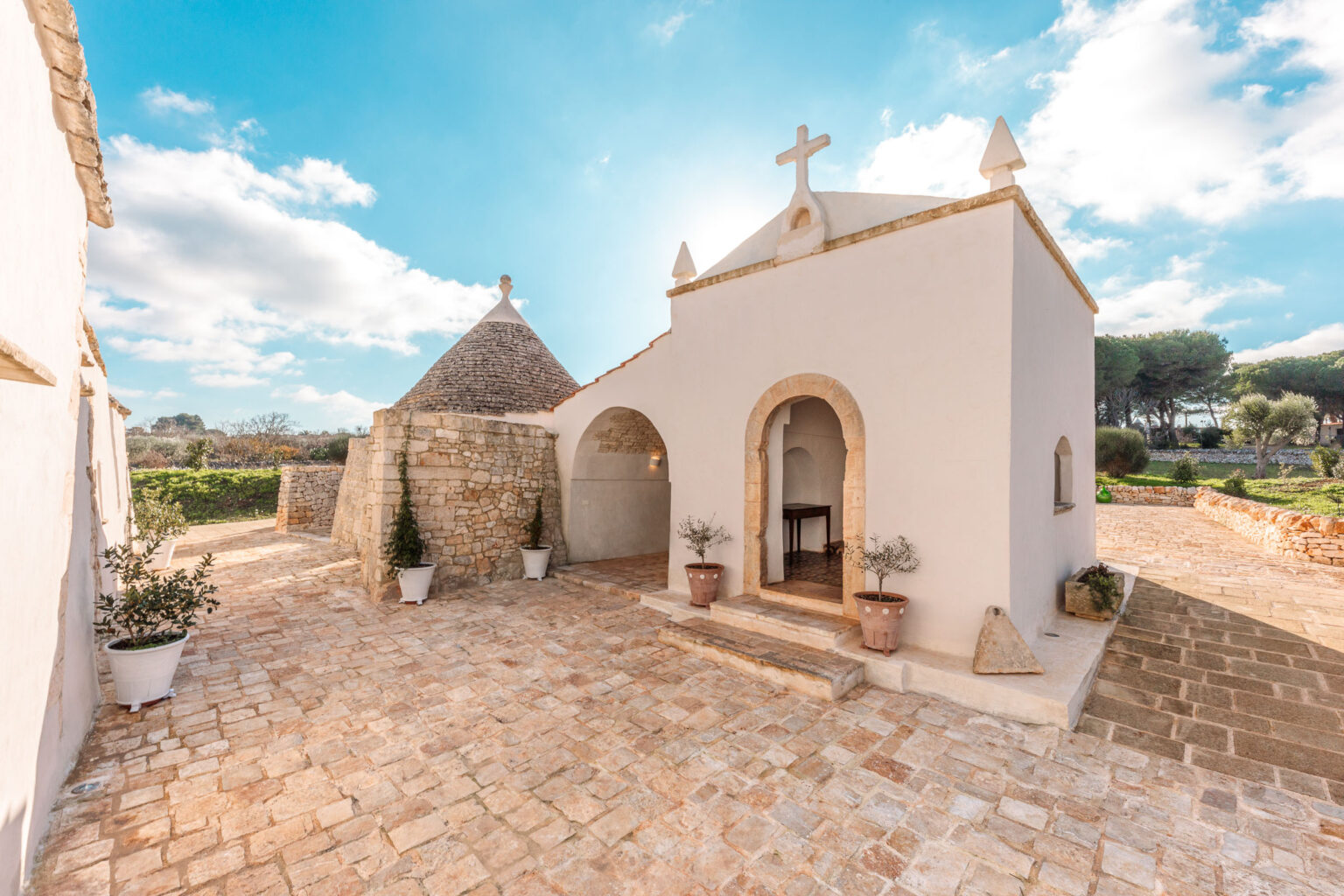 Charming white chapel with a conical stone roof, surrounded by a cobblestone pathway and potted plants, under a bright blue sky with scattered clouds. The setting features lush greenery in the background, highlighting the serene countryside atmosphere. | Only on SopranoVillas