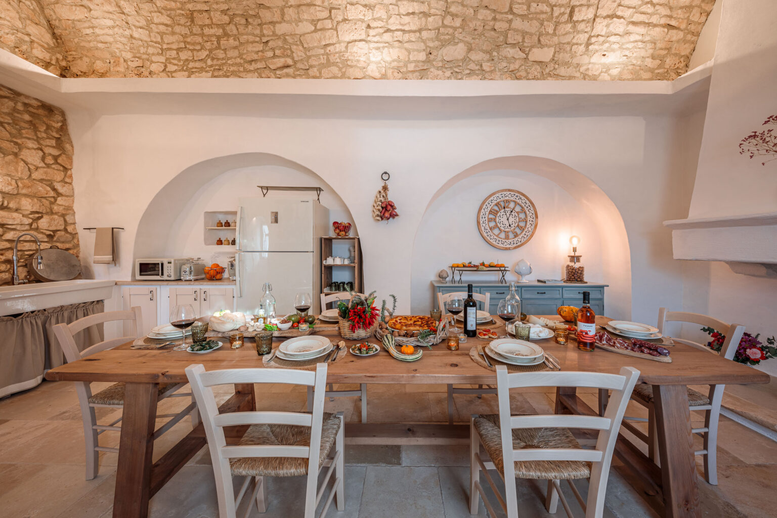 Modern kitchen dining area featuring a rustic wooden table set for a meal, surrounded by white chairs. The space has stone walls and arched doorways, with a cozy ambiance enhanced by decorative elements and fresh produce on the table. | Only on SopranoVillas