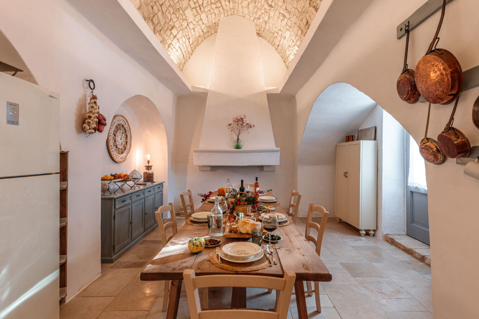 Cozy rustic kitchen featuring a wooden dining table set for a meal, with plates, glasses, and assorted foods. The space has arched ceilings, stone walls, and decorative elements like hanging pots and garlic. A sideboard holds fruits and a lamp, creating a warm and inviting atmosphere. | Only on SopranoVillas