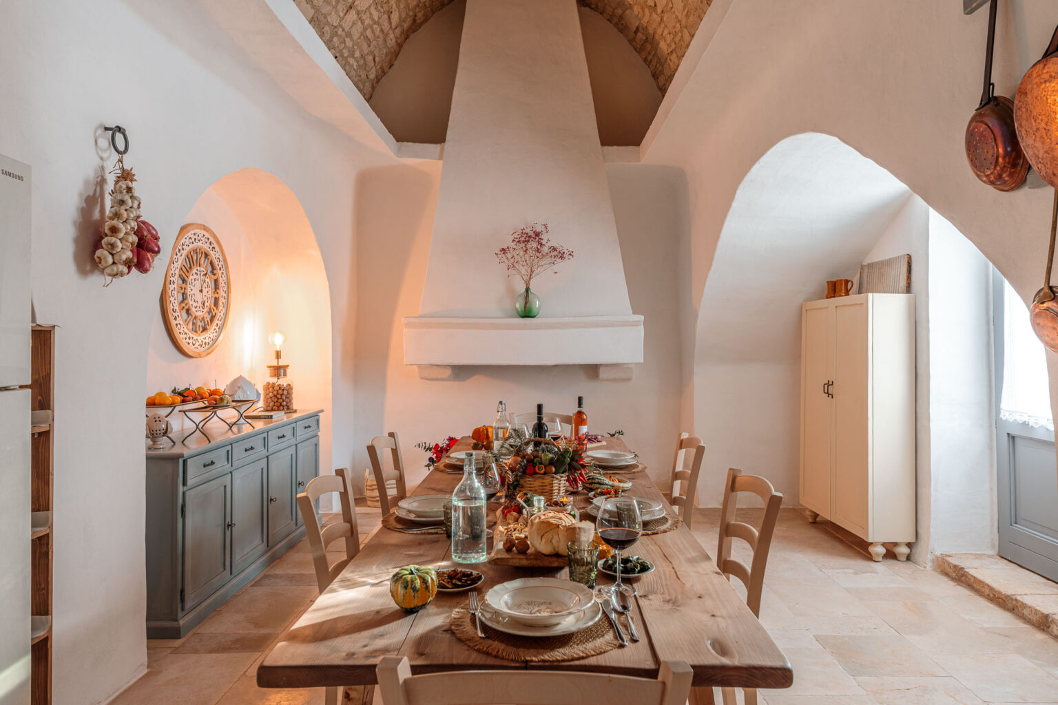 Cozy dining room with a wooden table set for a meal, featuring plates, glassware, and a centerpiece of fruits and vegetables. The room has a rustic charm with whitewashed walls, a vaulted ceiling, and decorative elements like a wall clock and hanging garlic. A sideboard displays additional food items, creating an inviting atmosphere for gatherings. | Only on SopranoVillas