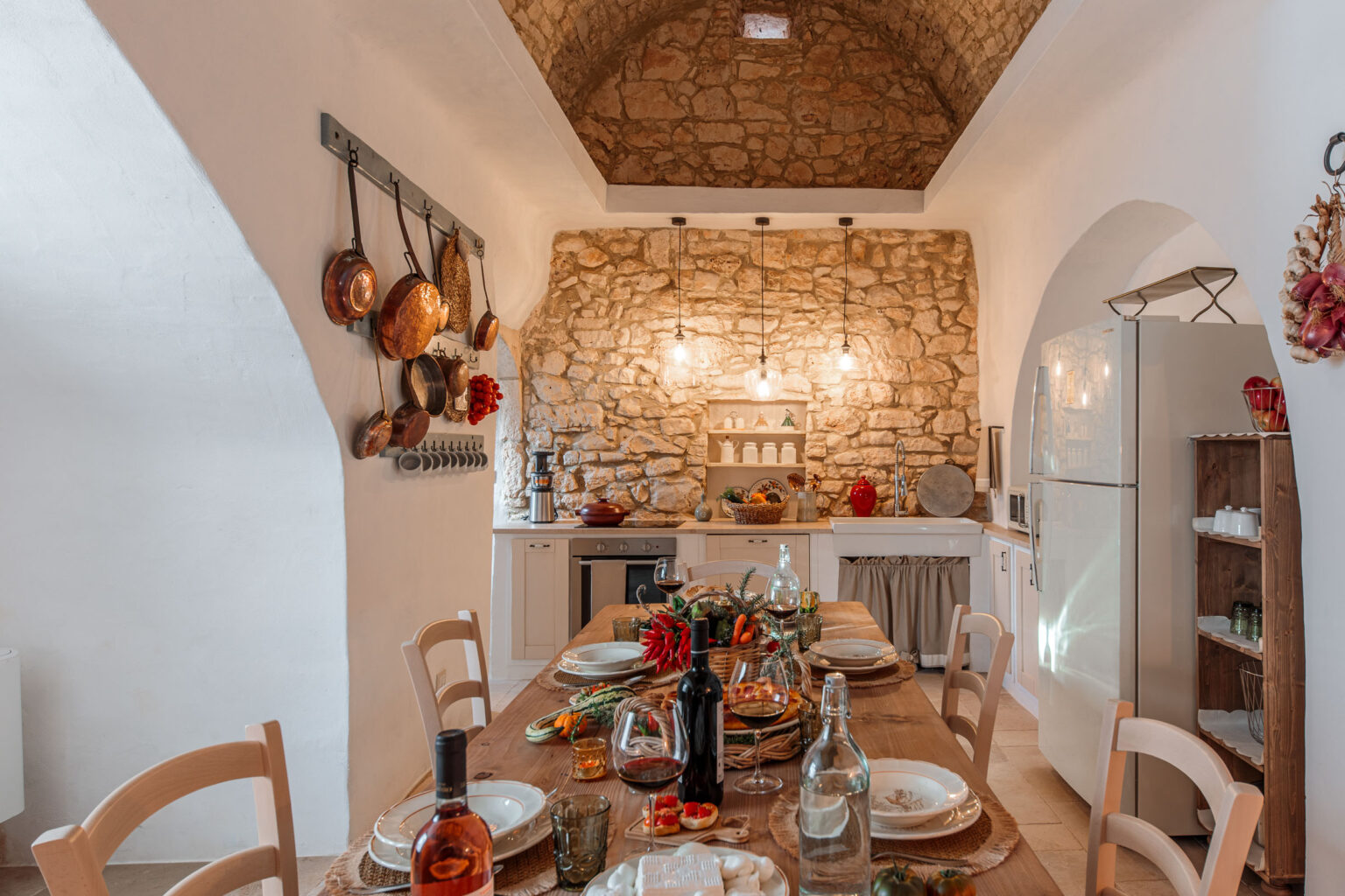 Rustic kitchen interior featuring a wooden dining table set with plates and glasses, surrounded by a stone wall and arched ceilings. Copper pots hang on the wall, with a modern refrigerator and shelves displaying kitchenware in the background. Warm lighting creates an inviting atmosphere. | Only on SopranoVillas