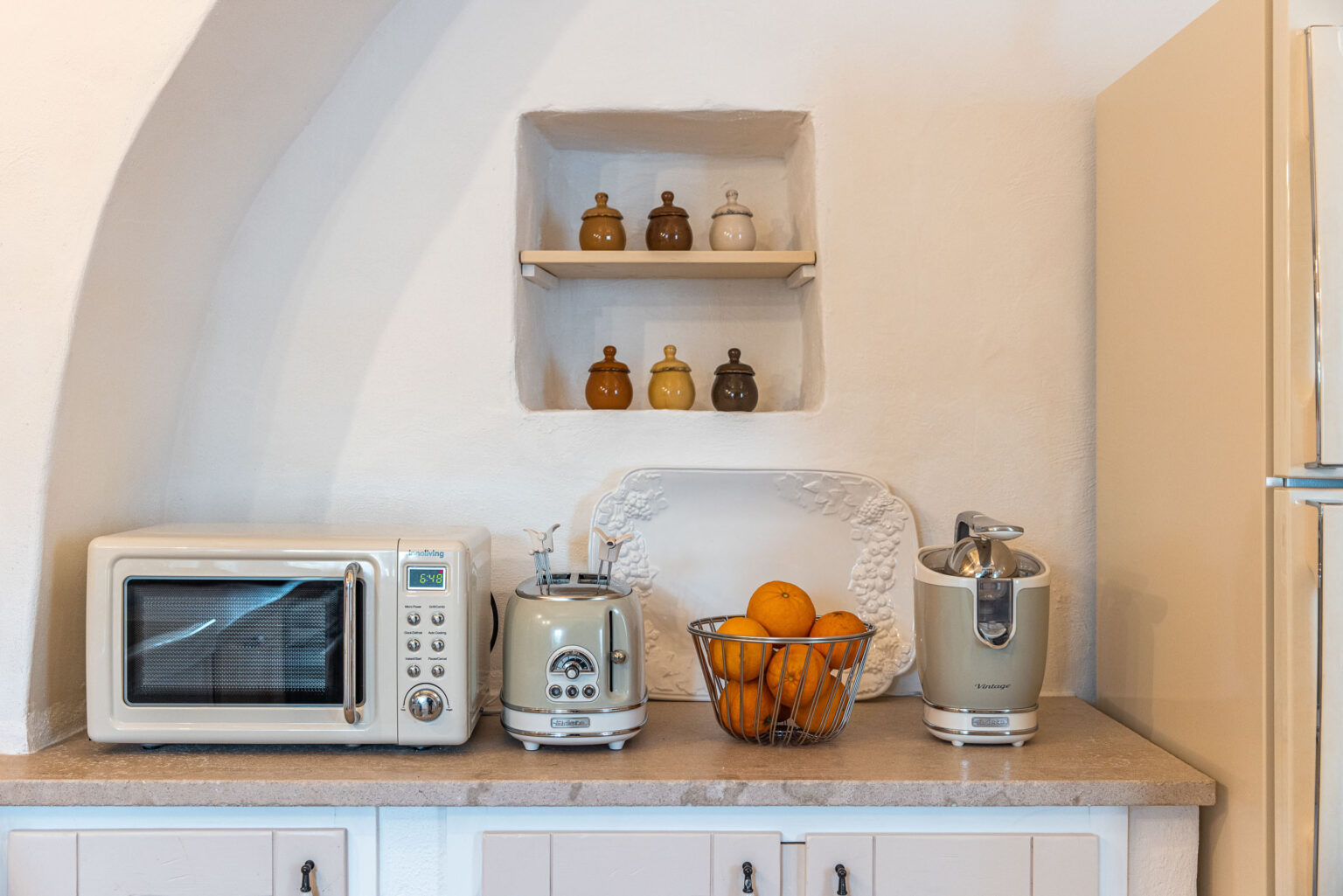 Modern kitchen countertop featuring a microwave, a vintage-style toaster, a bowl of oranges, and a juicer, with decorative jars on a shelf above. | Only on SopranoVillas