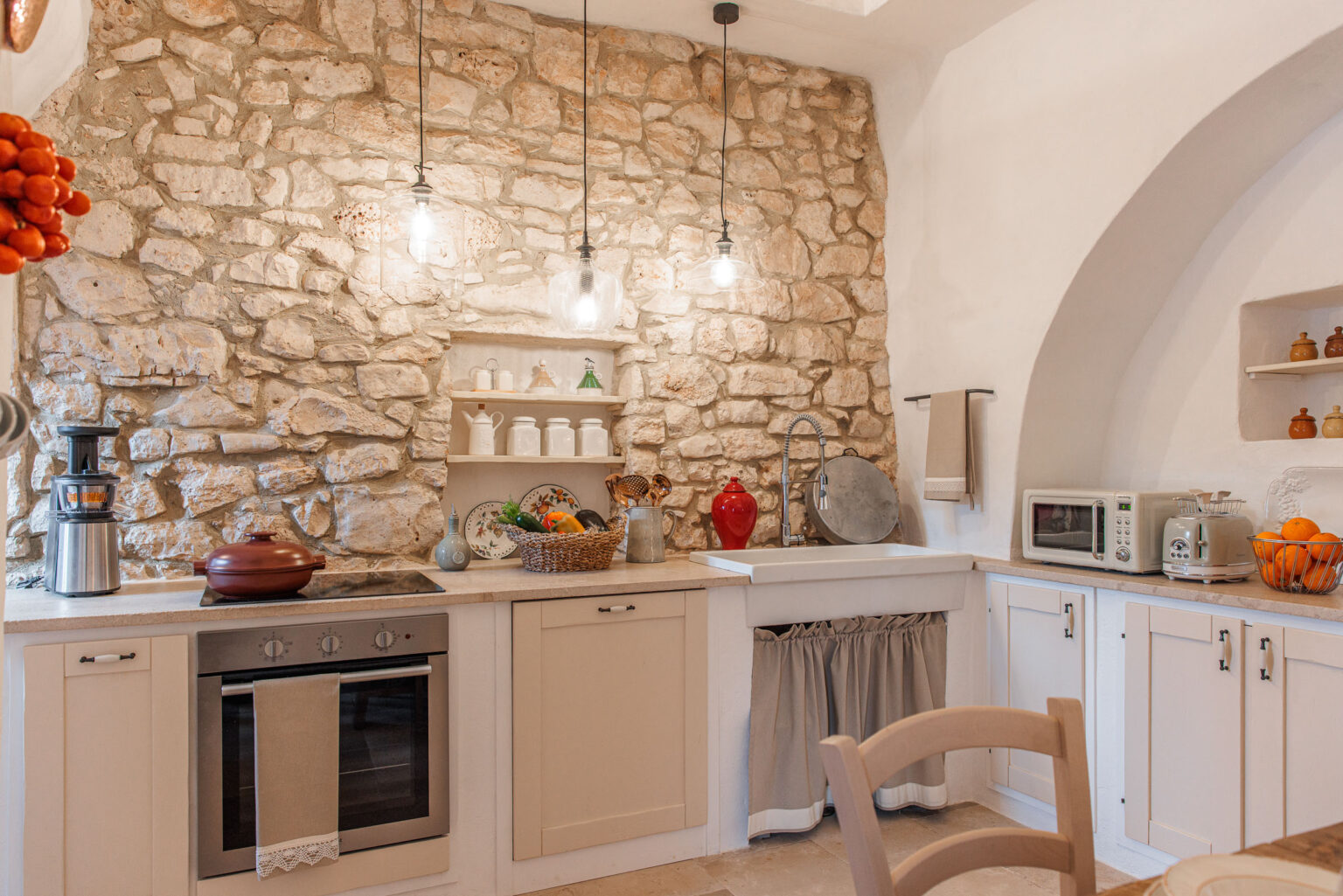 Cozy kitchen featuring a rustic stone wall, modern appliances, and wooden cabinetry. The space includes a large sink, stovetop, and decorative shelving displaying dishware and colorful fruits. Warm lighting enhances the inviting atmosphere, making it perfect for cooking and entertaining. | Only on SopranoVillas