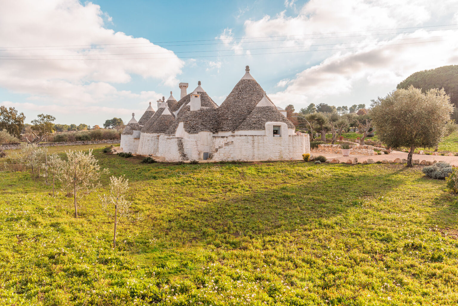 Trulli houses in the Italian countryside, surrounded by lush green grass and olive trees, under a partly cloudy sky. The unique conical roofs and whitewashed walls of the trulli showcase traditional Apulian architecture. | Only on SopranoVillas
