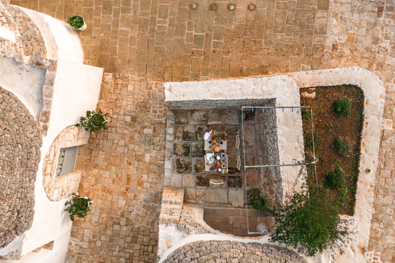Aerial view of a charming outdoor dining area surrounded by stone pathways and traditional architecture, featuring a table set with a variety of dishes and fresh vegetables, complemented by potted plants. | Only on SopranoVillas