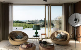 Modern living room with curved sofas, decorative pillows, and stylish accent tables. Large glass doors open to a scenic view of rolling hills and vineyards, enhancing the airy ambiance. A green lamp and a unique floor lamp add a touch of elegance to the interior design.