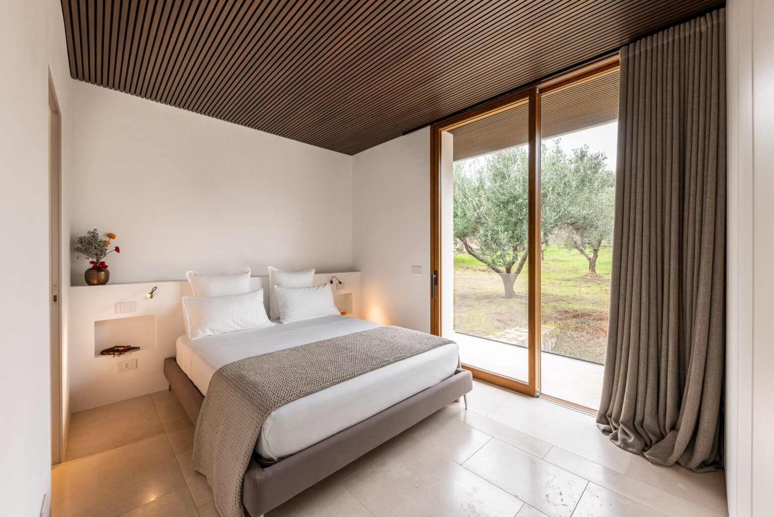 Modern bedroom featuring a double bed with gray bedding, large windows with natural light, and minimalist decor. The room has wooden ceiling accents and a view of olive trees outside. | Only on SopranoVillas